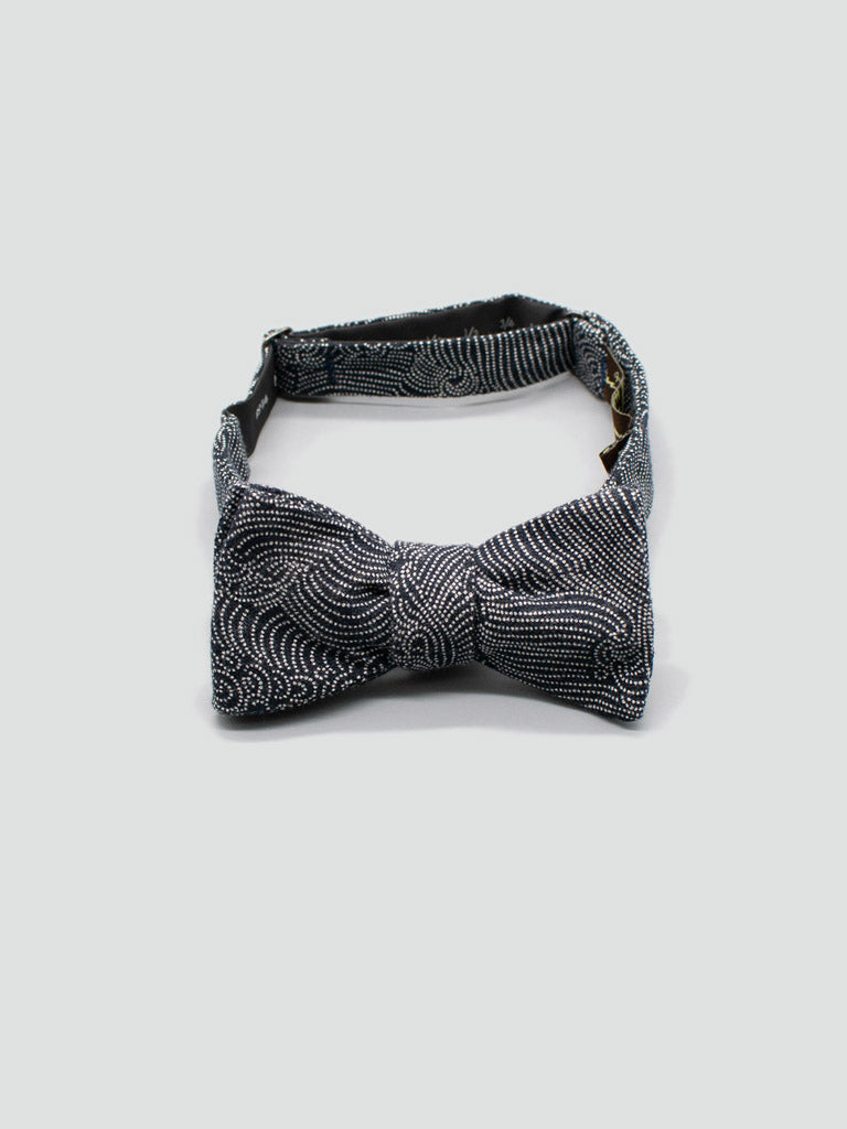 The Japanese Nami Printed Bow Tie by ID Menswear features an intricate black and white swirl pattern and comes with an adjustable strap and metal clasp. Handmade in Brooklyn, its displayed on a plain light gray background.