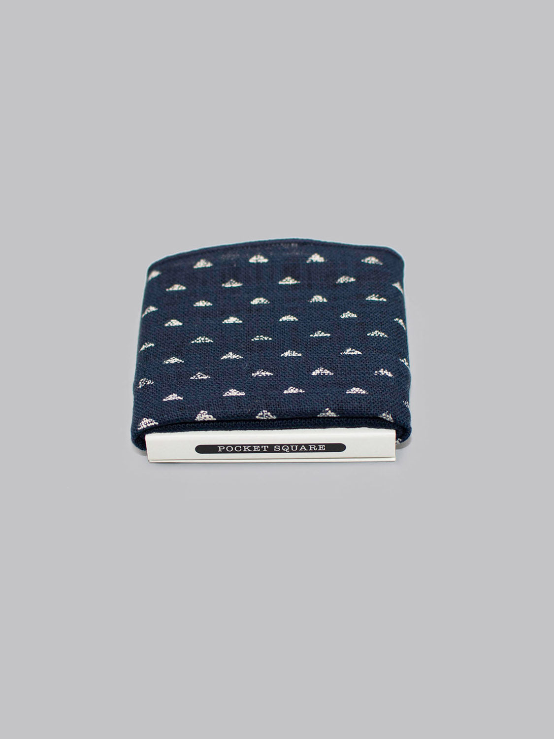 The Nara Pocket Square by ID Menswear, a navy blue cotton piece with a small white pattern, is handmade in Brooklyn and shown neatly folded with a silver POCKET SQUARE clip against a light gray background.