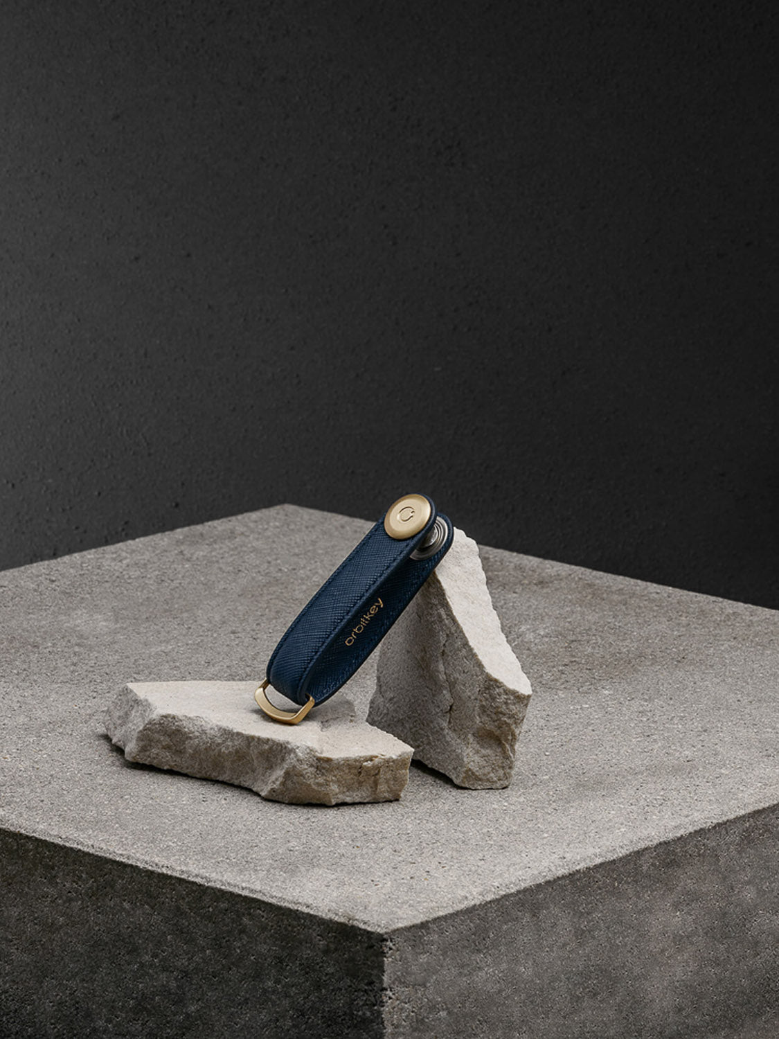 The Orbitkeys Key Organizer Saffiano Leather, featuring a navy strap and gold hardware, stands upright on two broken concrete slabs against a textured gray backdrop.
