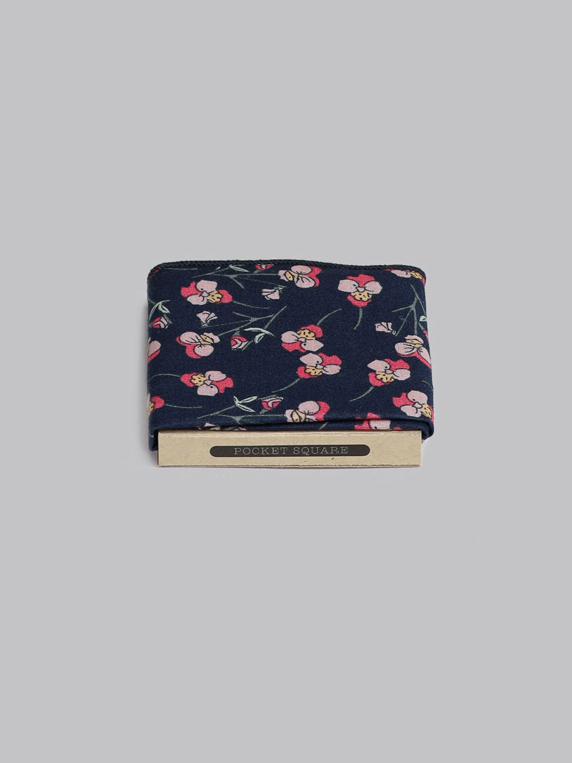 The Navy Pop Pocket Square by ID Menswear features a pink and white floral pattern on 100% cotton, handmade in Brooklyn. It is neatly folded in a beige holder labeled POCKET SQUARE, set against a plain gray background.