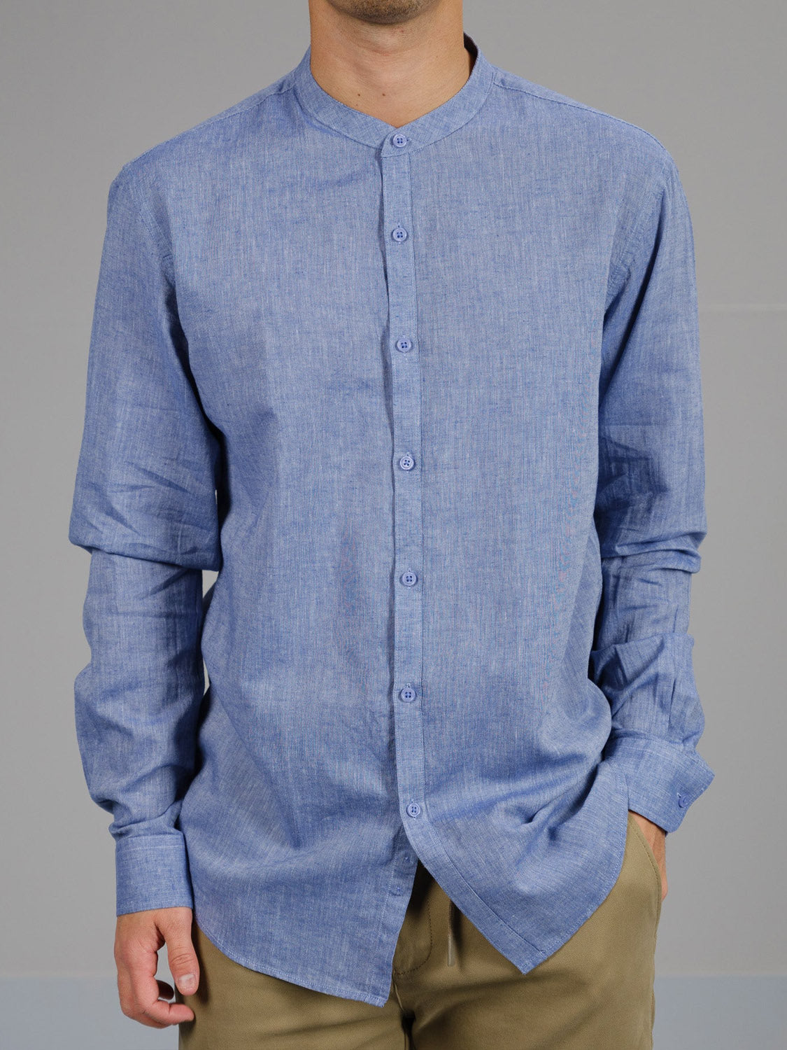 A person wears an untucked blue ID Menswear Nehru Linen Cotton Mandarin Collar Shirt with light brown pants, resting their left hand casually in the pocket against a plain gray background.