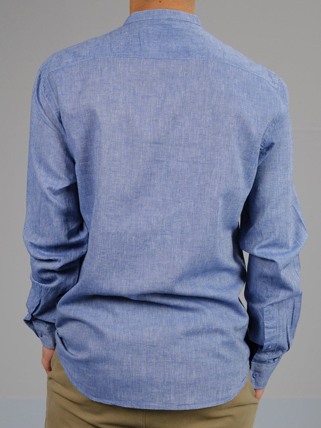 A person wearing the Nehru Linen Cotton Mandarin Collar Shirt by ID Menswear—a light blue, breathable linen-cotton blend—stands with their back to the viewer in beige pants against a plain grey background, adding a minimalist touch.