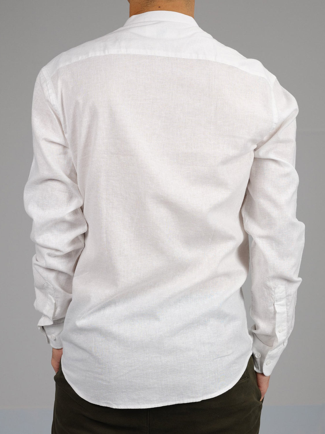Back view of a person in the Nehru Linen Cotton Mandarin Collar Shirt by ID Menswear, featuring a breathable linen cotton blend and buttoned cuffs. The plain white shirt is paired with dark pants against a neutral gray background.