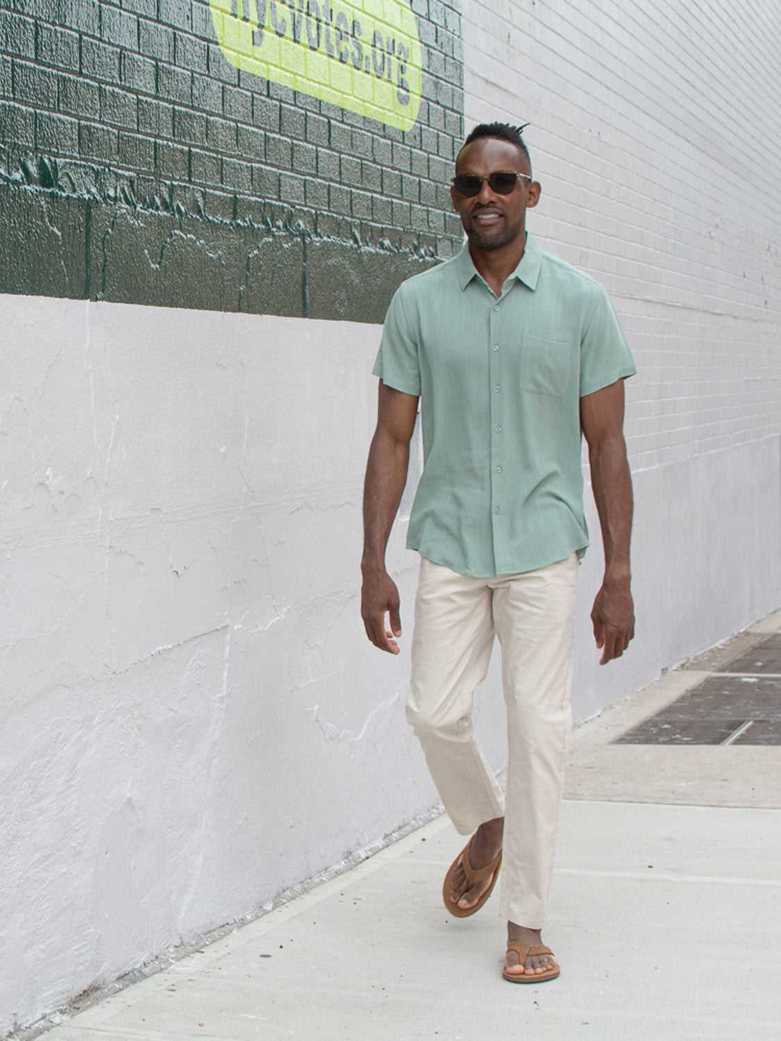 A man in sunglasses, cream pants, brown sandals, and the New Hampton Linen Blend Shirt by ID Menswear walks on a sidewalk past a white and green brick wall.