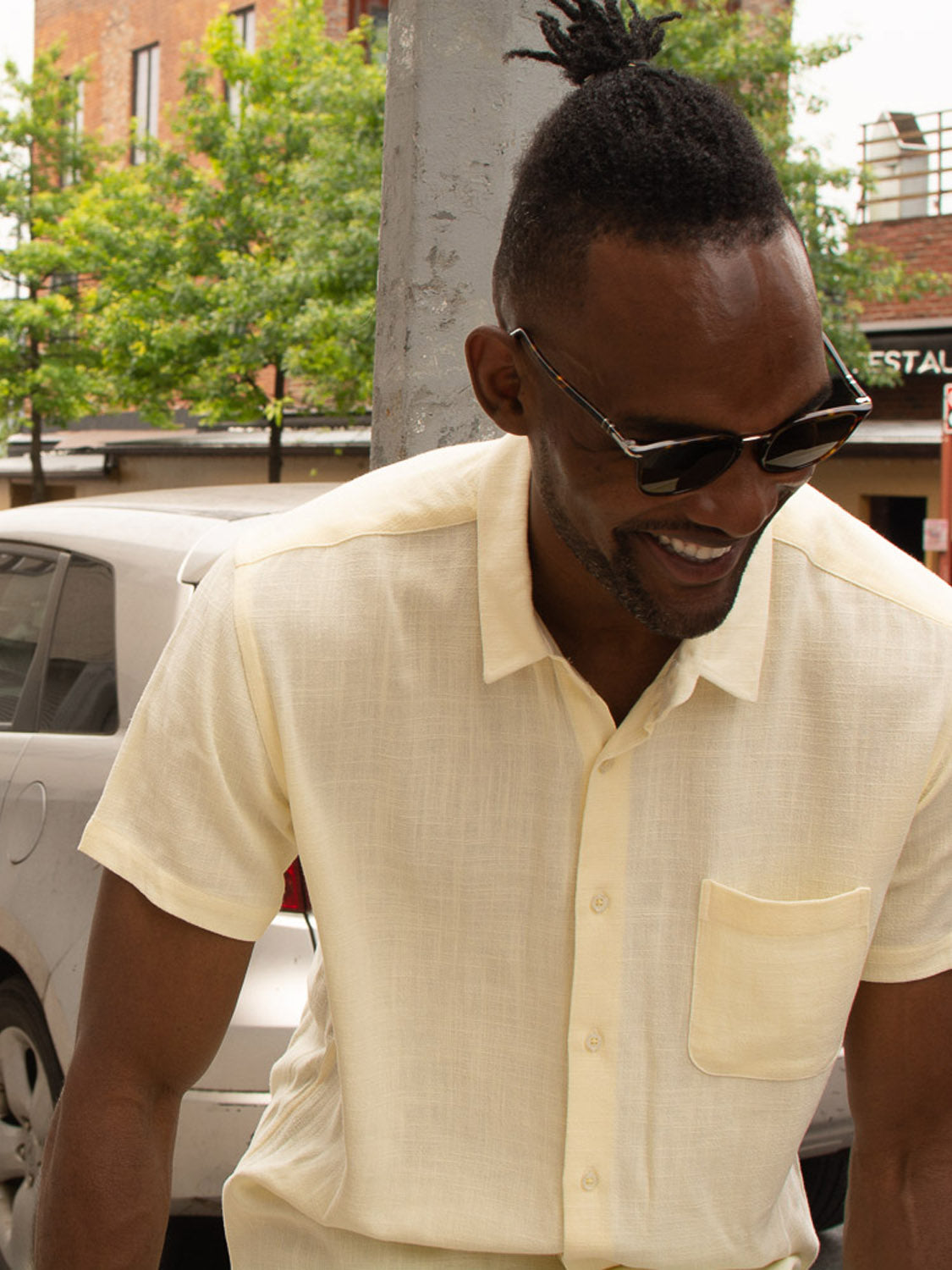 A man in sunglasses and the ID Menswear New Hampton Linen Blend Shirt smiles while standing outdoors near a parked car and leafy trees.
