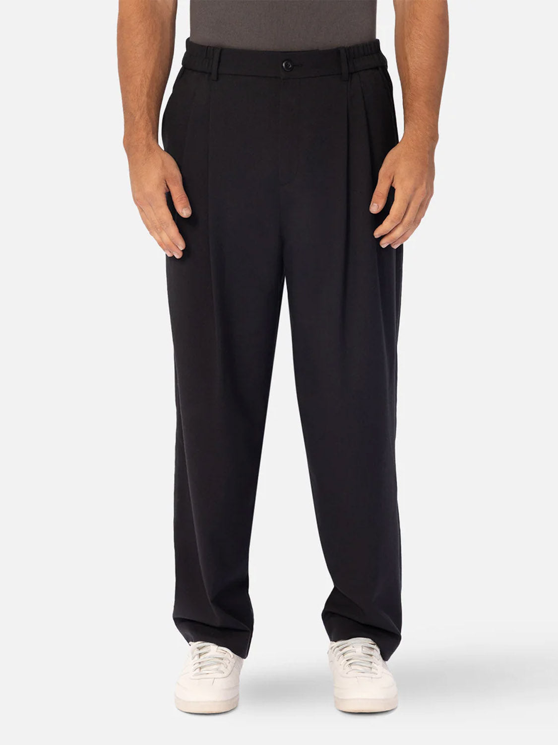 A person stands against a plain light background wearing Industrie’s The New Milano Pants - Loose Fit in a smart casual style, paired with a tucked-in dark shirt and white sneakers, highlighting the pants’ relaxed silhouette from shoulder to hem.