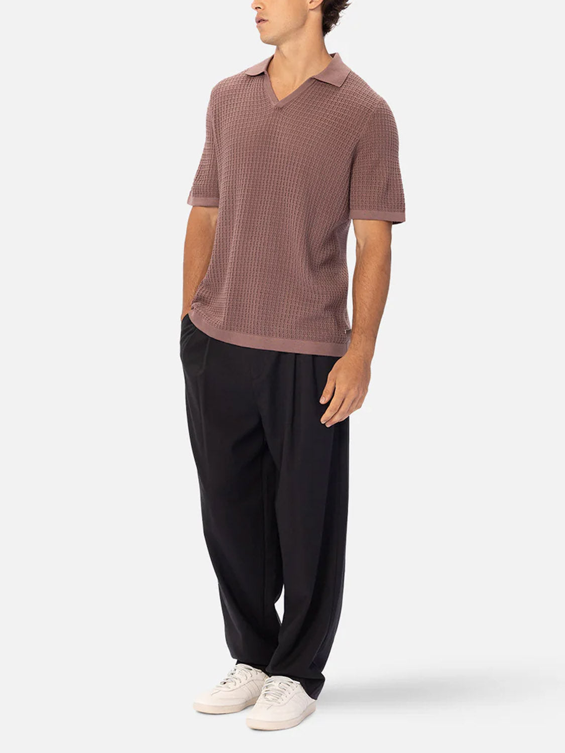 A man stands with his hands in his pockets, wearing a textured mauve short-sleeve polo, Industrie’s The New Milano Pants - Loose Fit, and white sneakers against a plain light background.