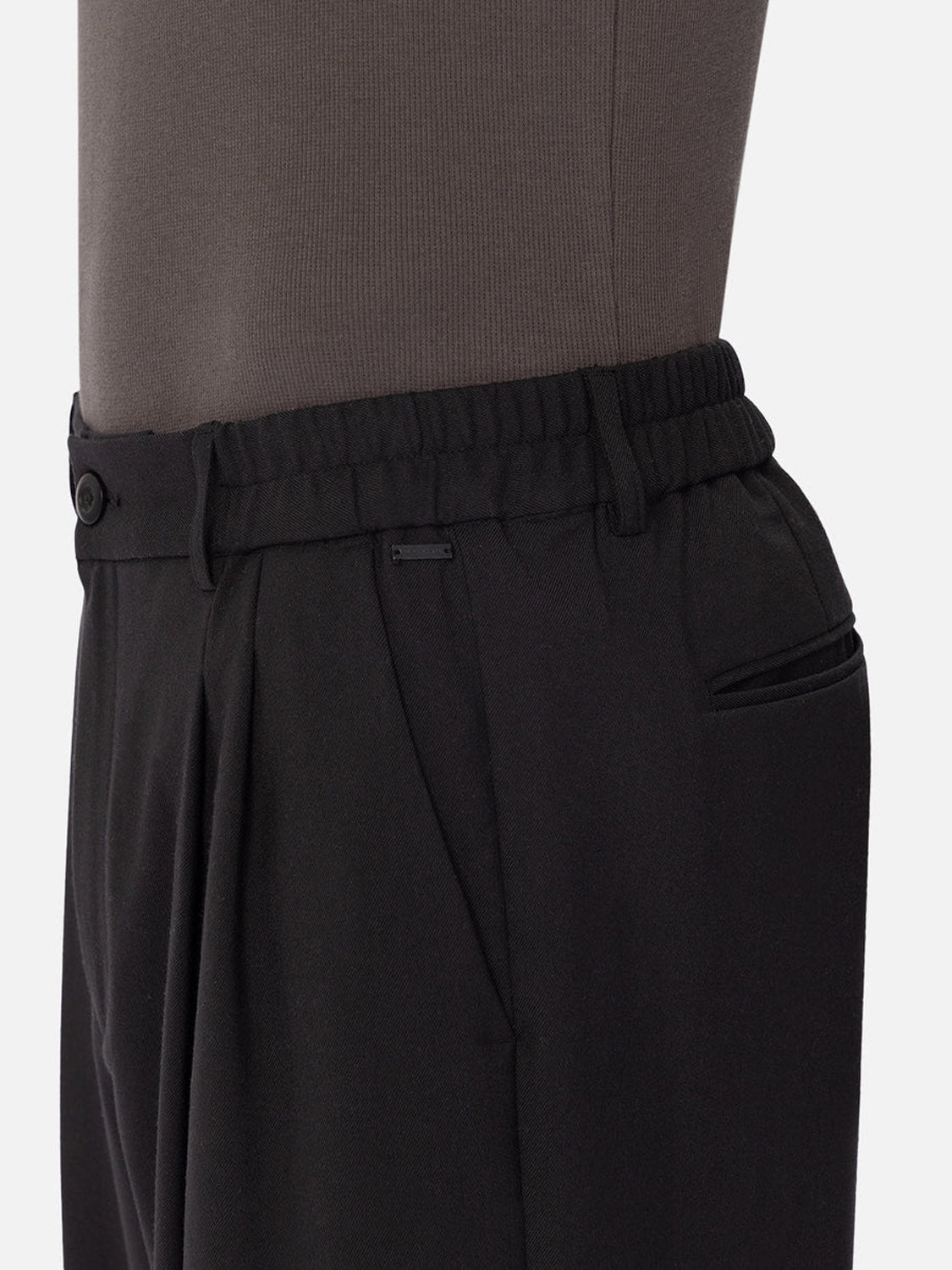 A close-up side view shows a person wearing Industrie’s The New Milano Pants - Loose Fit, featuring an elastic waistband, single button closure, pleats, and a side pocket, paired with a tucked-in dark top.