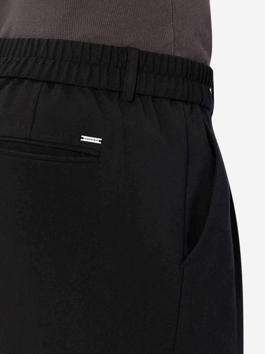 A close-up of a person wearing Industrie’s The New Milano Pants - Loose Fit in black, featuring an elastic waistband, side pocket, and a small metallic label above the back pocket, paired with a dark grey top for a smart casual look.