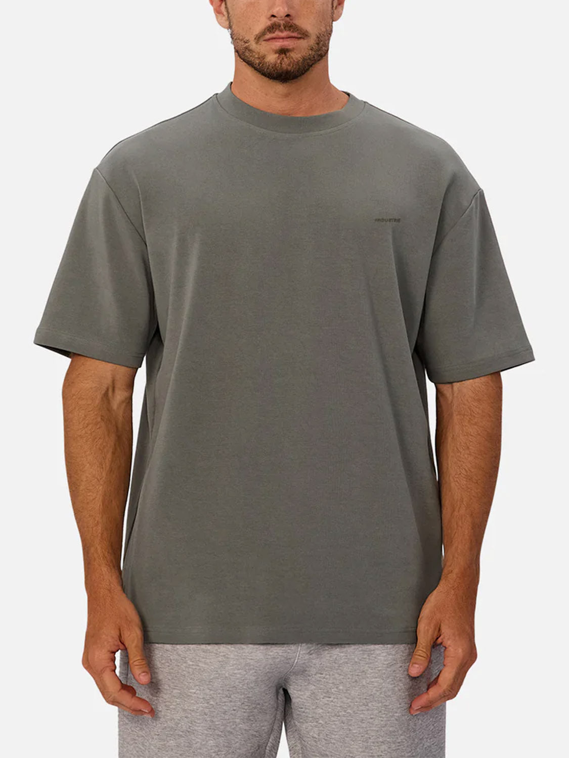 A man stands facing forward against a plain light background, wearing Industrie’s The Newport T-Shirt—an oversized, heavyweight grey 90s-inspired style—paired with light grey shorts.