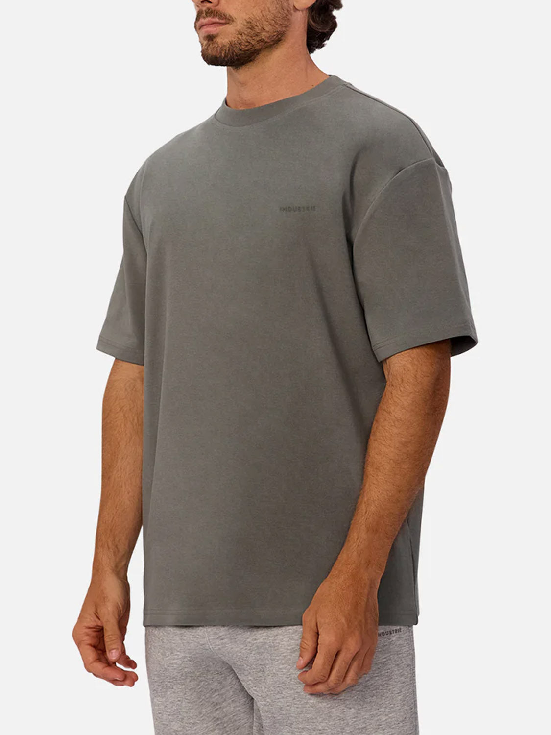 A man stands against a plain white background, facing slightly to the side, wearing Industrie’s The Newport T-Shirt—an oversized 90s-inspired gray tee—paired with light gray shorts.