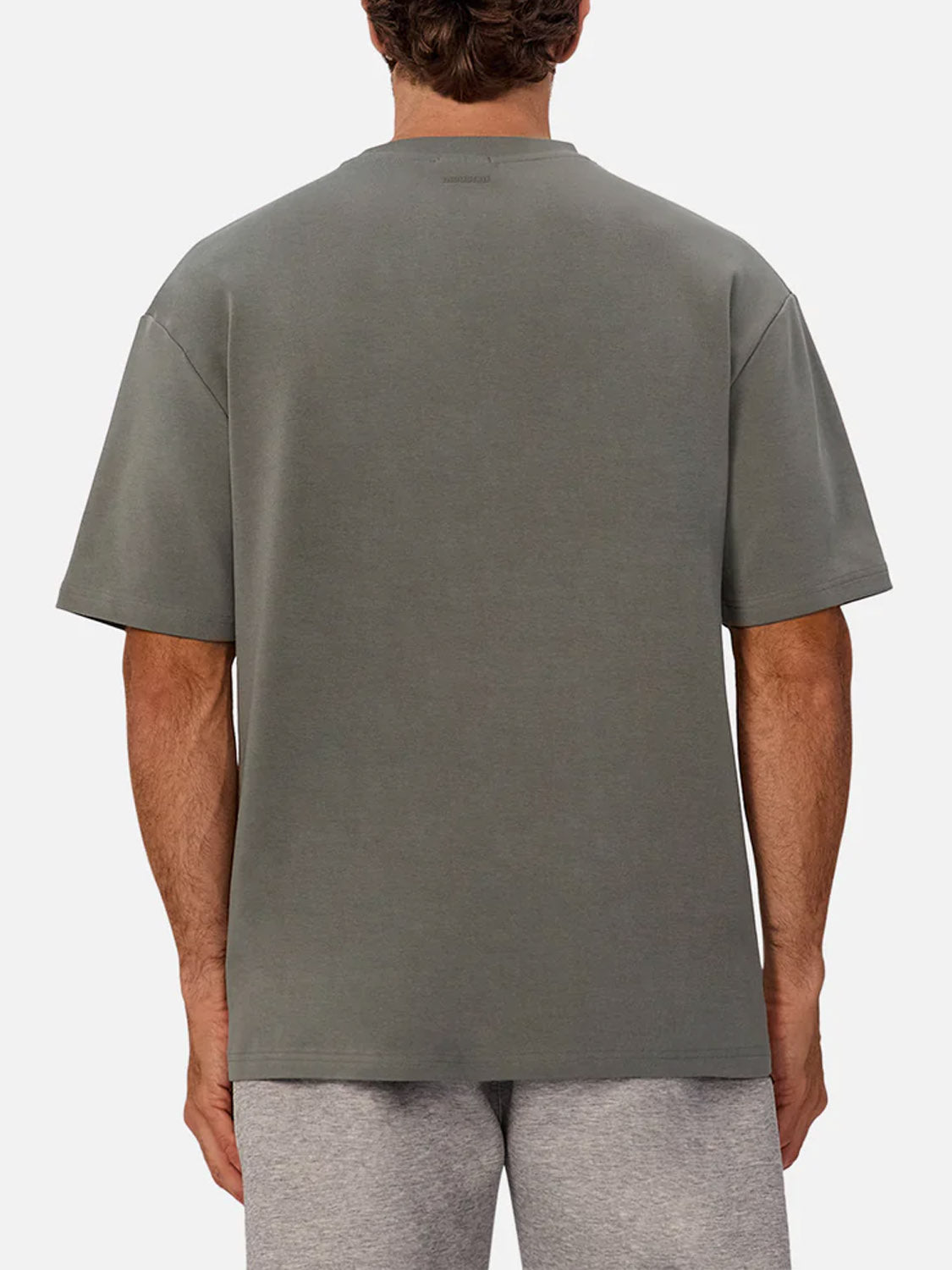 A person is seen from behind wearing The Newport T-Shirt by Industrie, a 90s-inspired oversized grayish-green tee, paired with light gray shorts against a plain, light background.