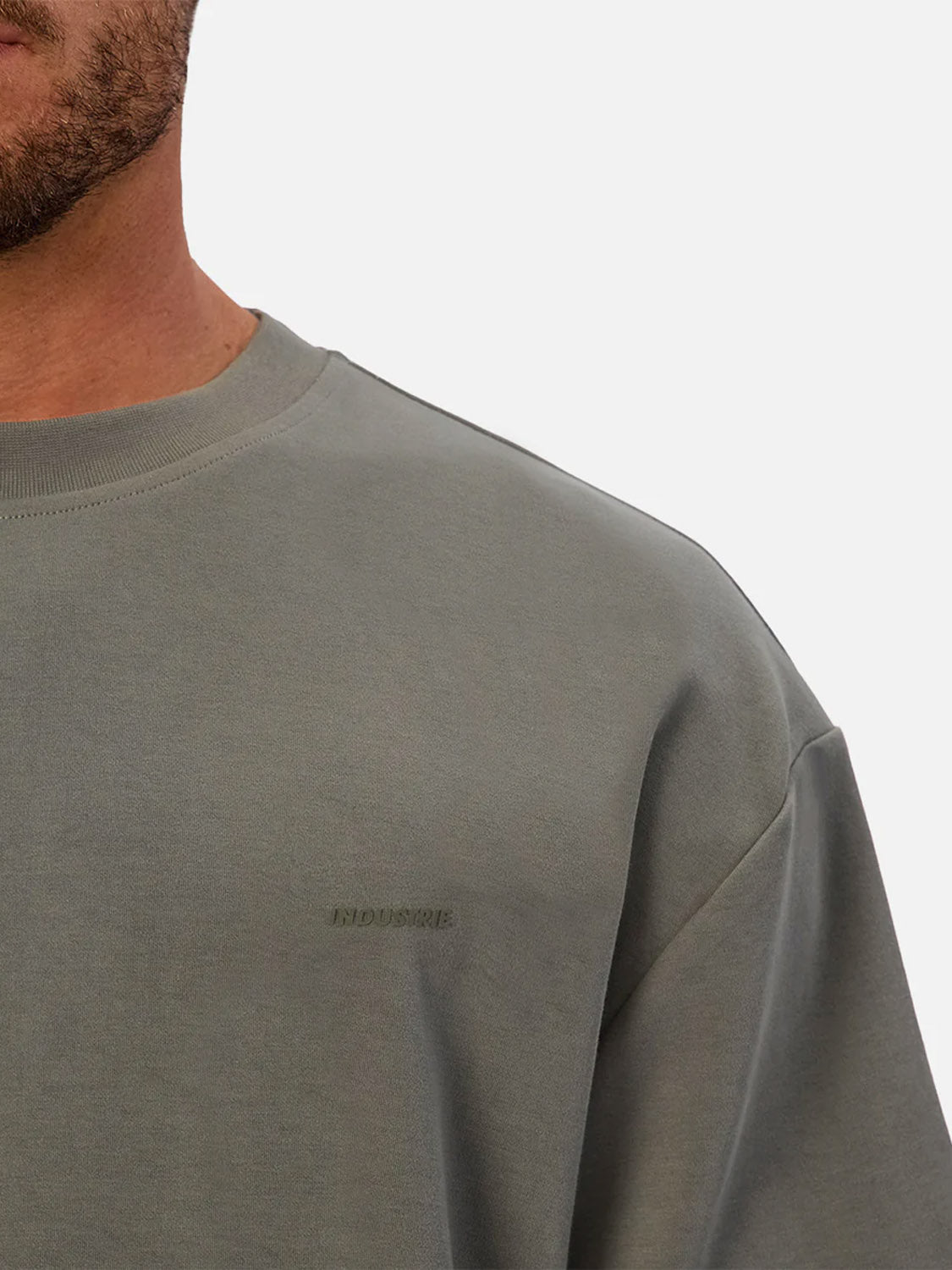 A close-up shows a person wearing The Newport T-Shirt by Industrie, a gray, 90s-inspired crew-neck with subtle chest embroidery. Only the lower face, neck, and upper torso appear against a plain background.