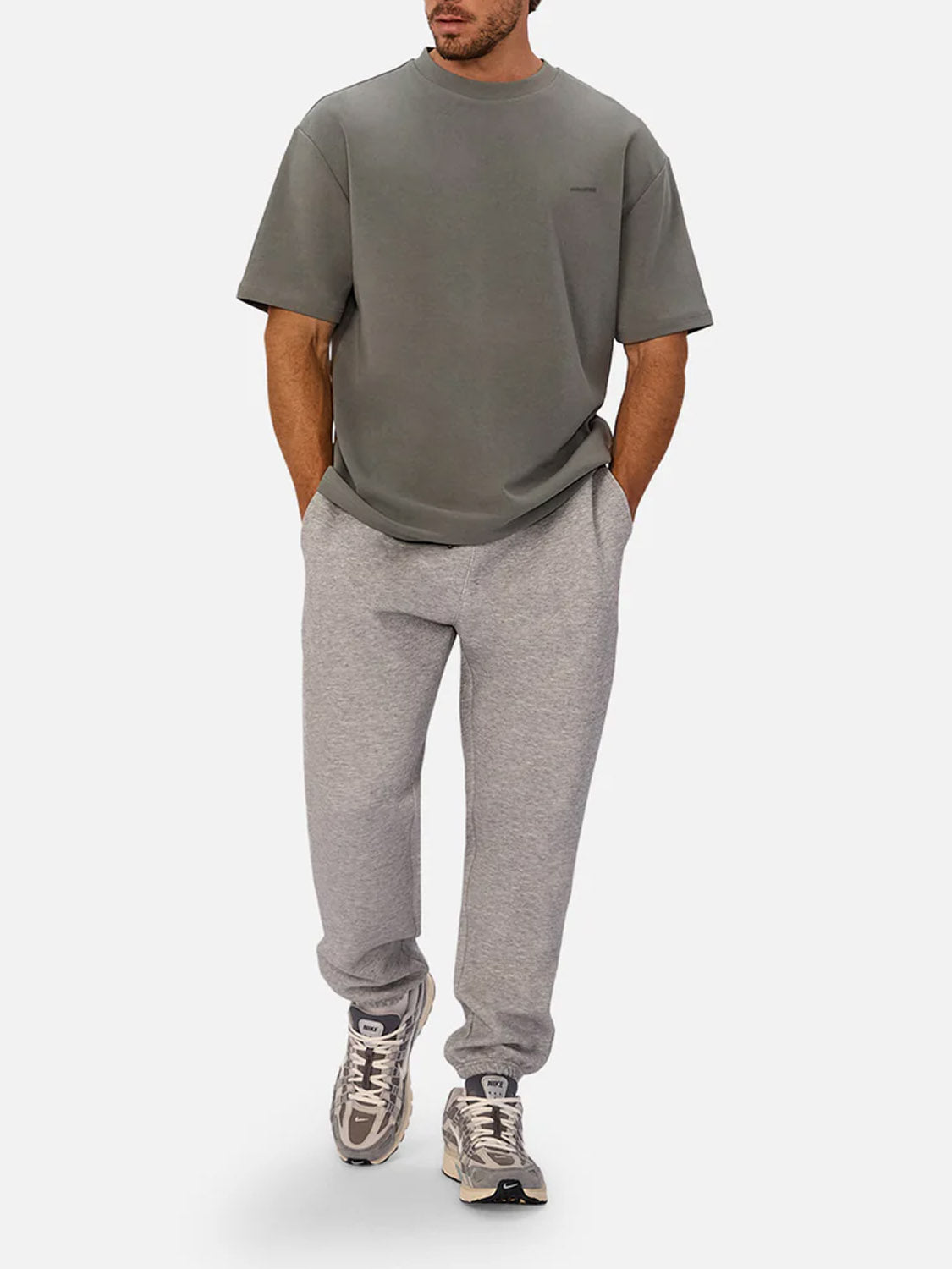 A man stands with his hands in his pockets against a plain white background, wearing Industrie’s The Newport T-Shirt—an oversized, 90s-inspired gray heavyweight tee—paired with light gray sweatpants and gray sneakers with white accents.