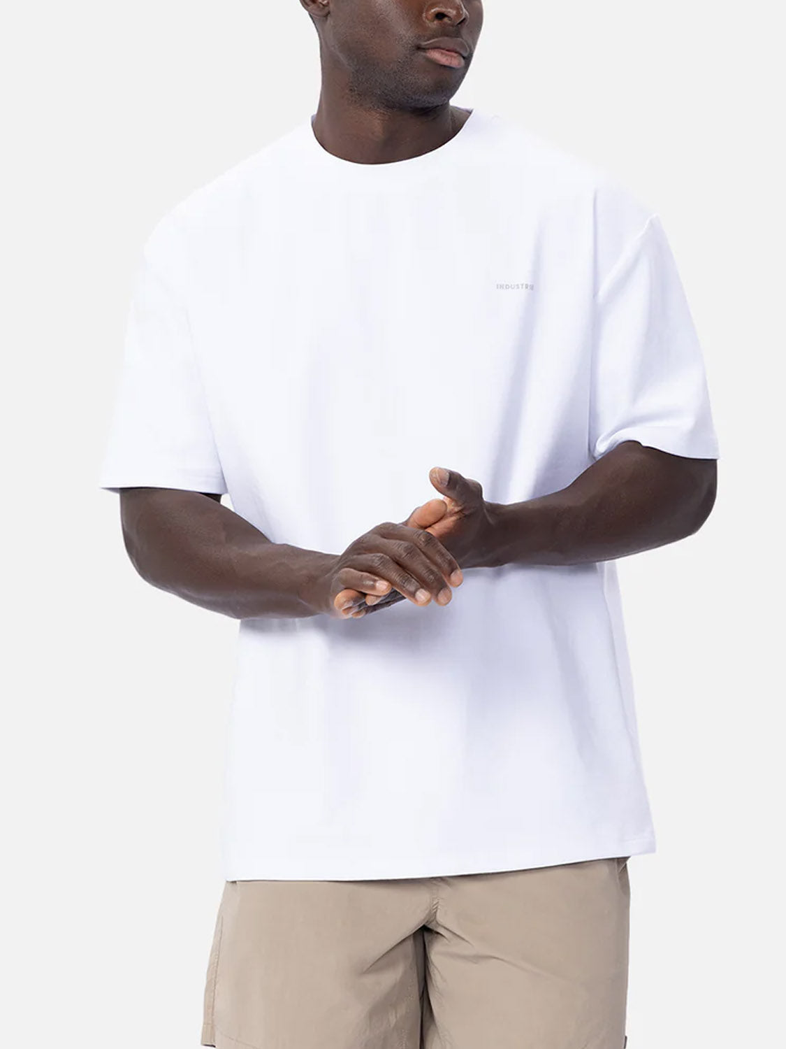 A man wears The Newport T-Shirt by Industrie and beige shorts, standing against a light gray background with his hands clasped and looking to the side.