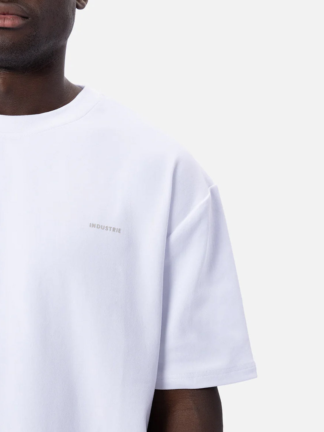 A person wears The Newport T-Shirt by Industrie—a 90s inspired, plain white oversized tee with a subtle INDUSTRIE chest print—shown from the shoulders down against a light gray background.