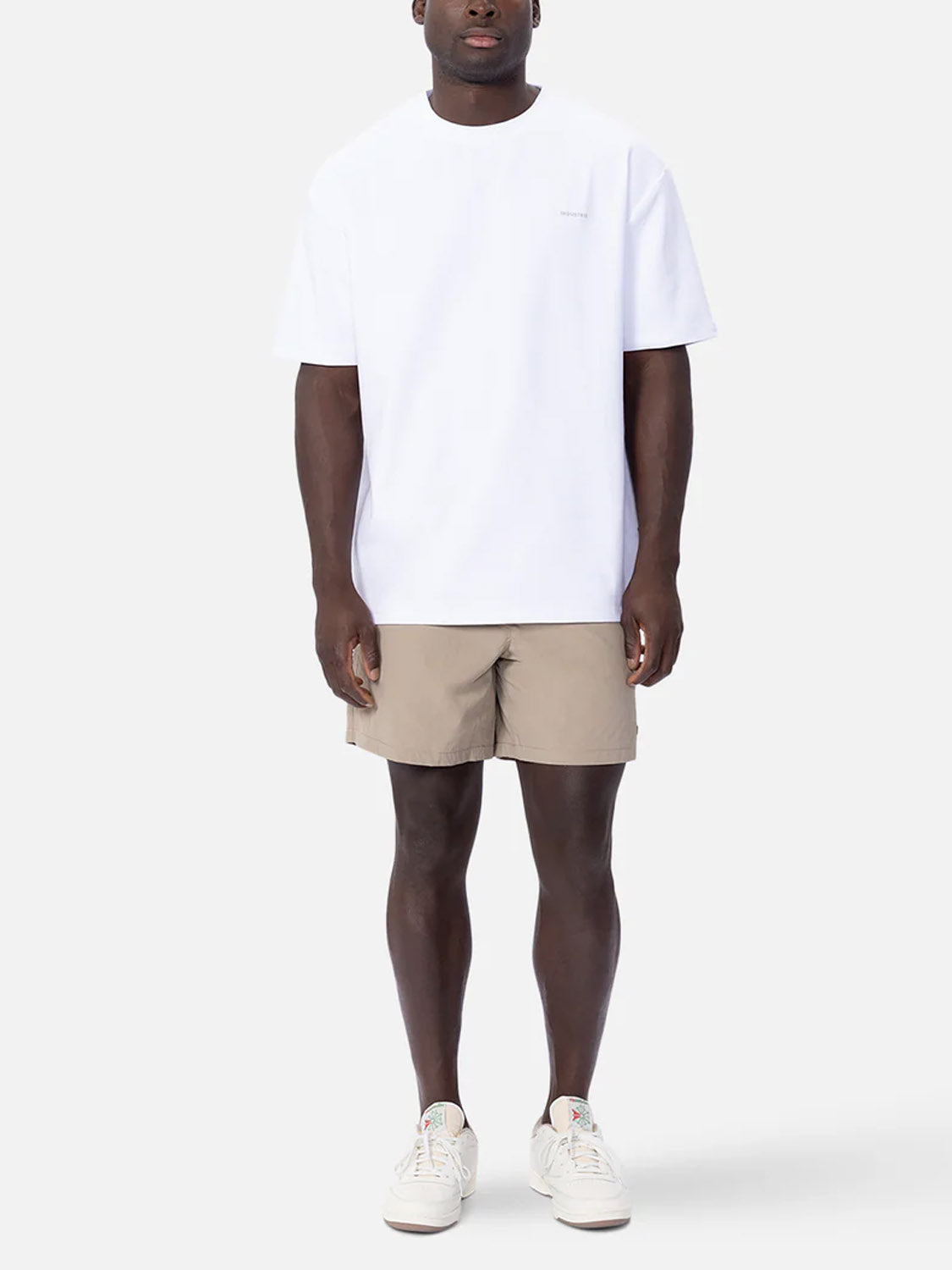 A person stands against a plain background wearing Industrie’s The Newport T-Shirt, paired with beige shorts and white sneakers for a casual, minimal look. They face forward with arms relaxed at their sides.