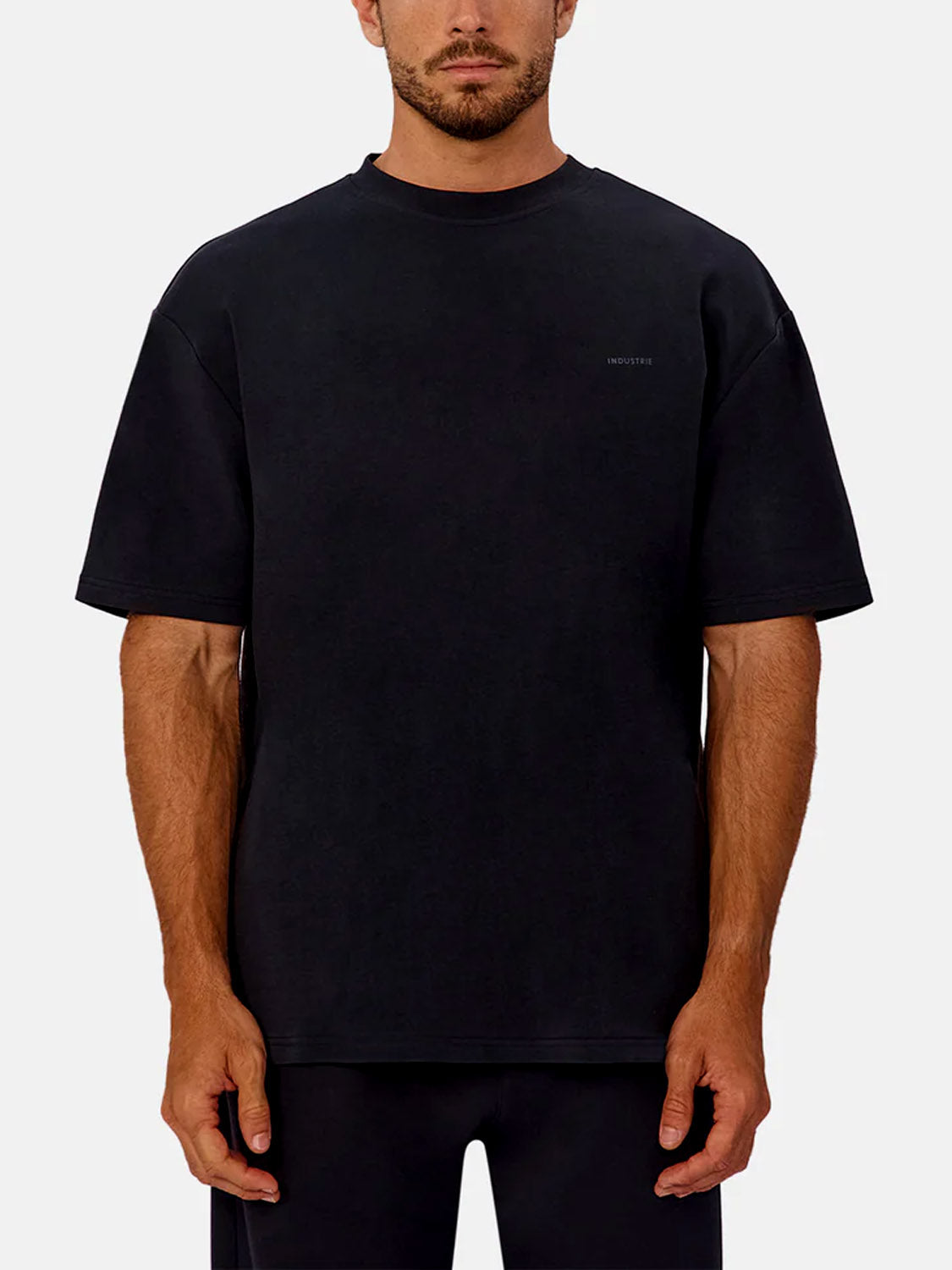 A man faces forward in a loose-fitting, heavyweight Industrie Newport T-Shirt in black with matching black pants, standing against a plain light grey background.