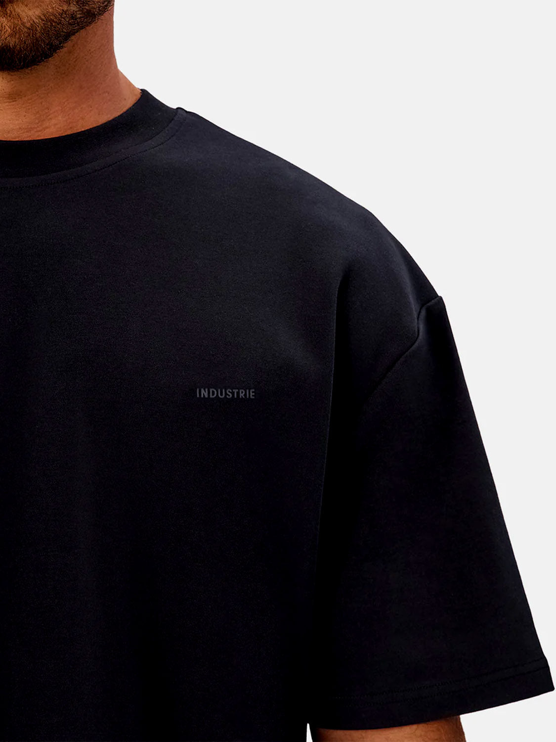 A person wearing The Newport T-Shirt by Industrie—a plain black, heavyweight crew neck tee with subtle INDUSTRIE lettering on the chest. Only the upper torso is shown against a light background.