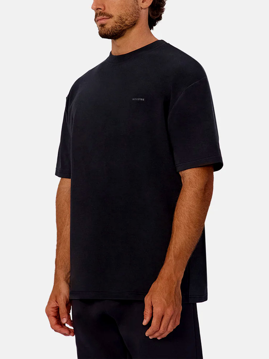 A man stands against a plain white background, turned slightly to the side, wearing Industrie’s The Newport T-Shirt—a black, oversized, heavyweight crew neck tee with short sleeves and subtle chest text.