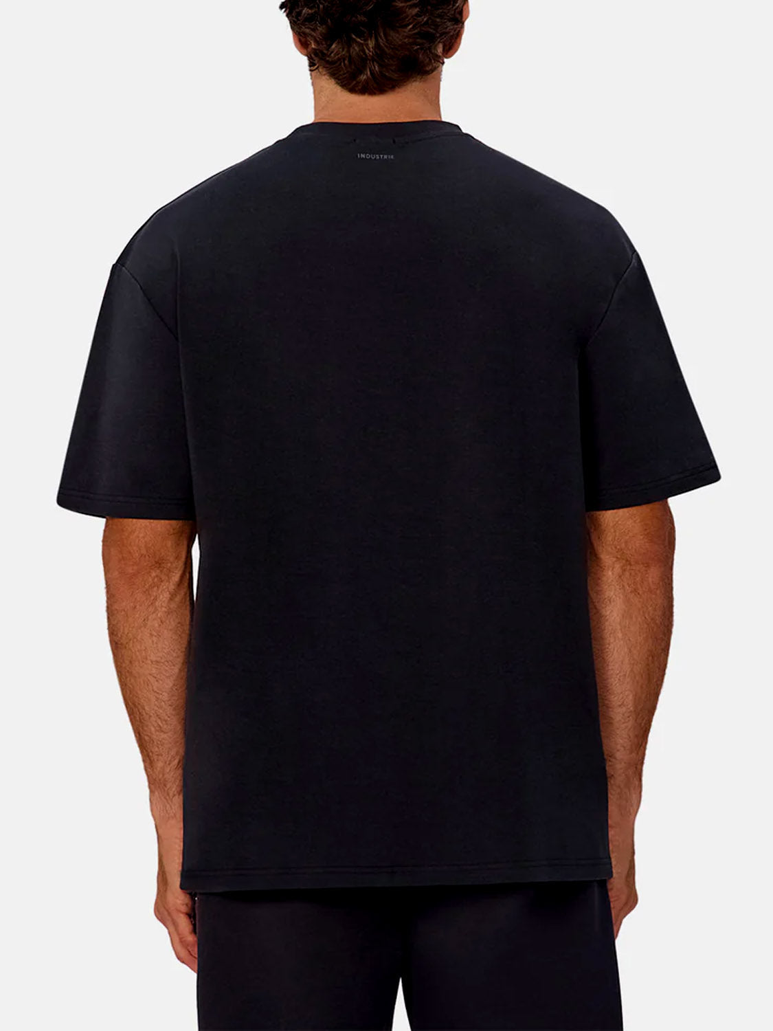 A man faces away from the camera in a loose-fitting Newport T-Shirt by Industrie and dark pants, set against a plain light gray background.