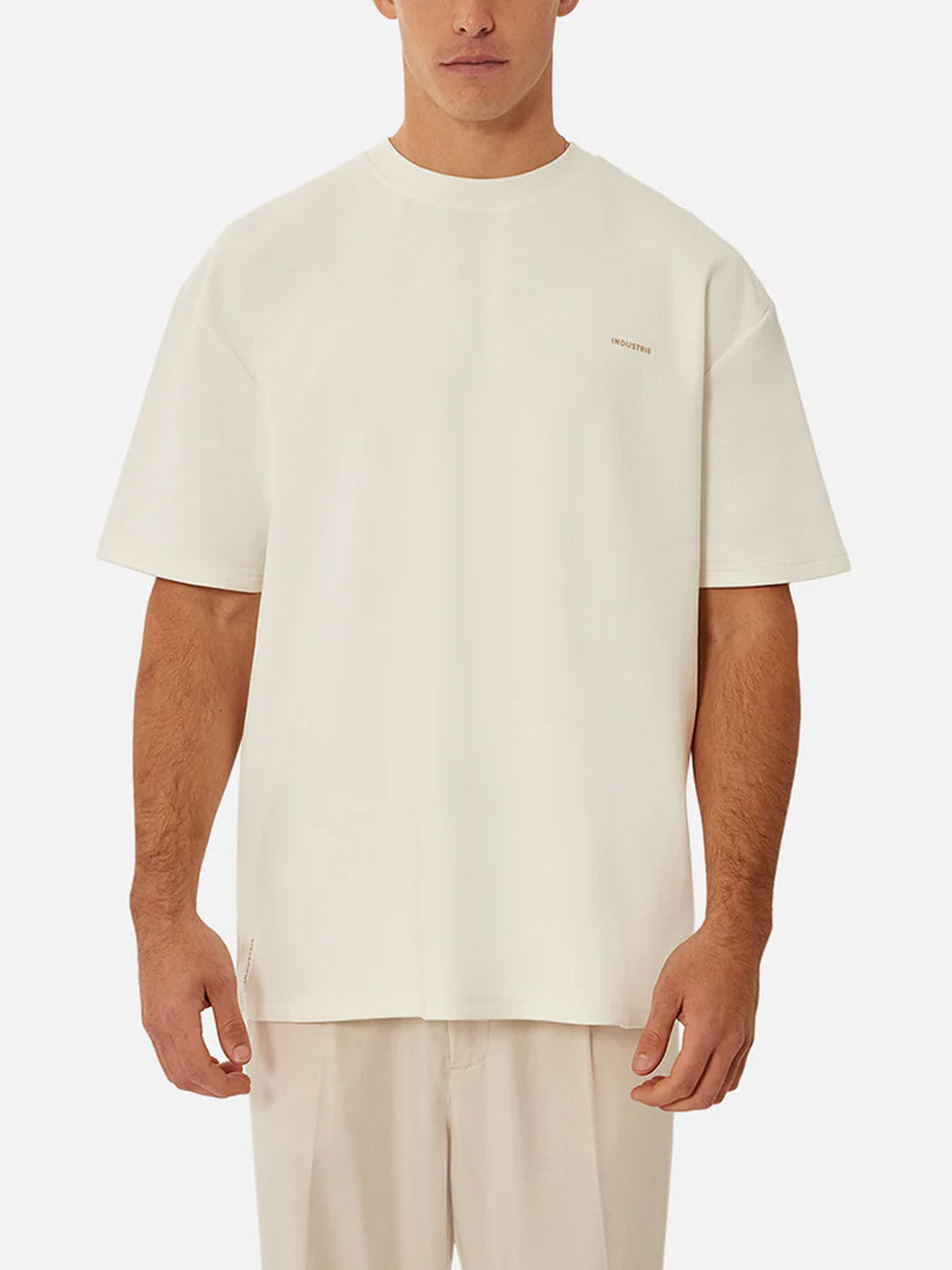 A man stands facing forward against a plain white background, wearing Industrie’s The Newport T-Shirt—an oversized, off-white heavyweight crew neck—paired with matching light-colored pants.