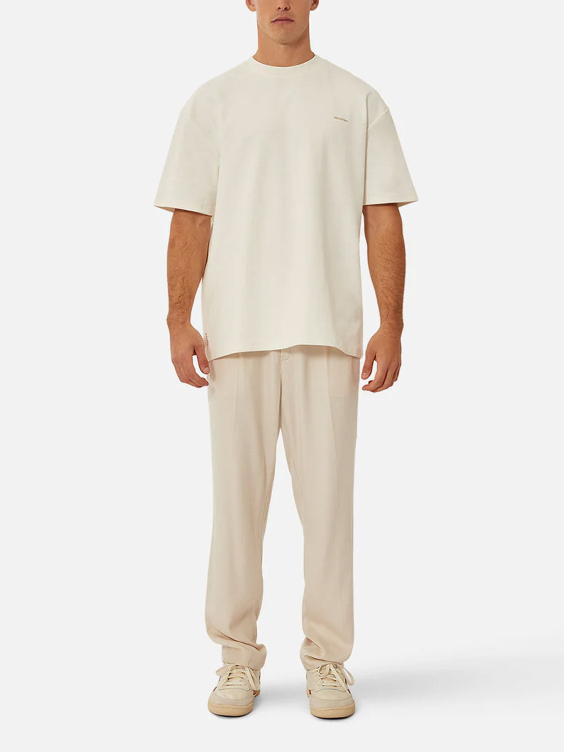 A man faces forward against a plain white background, wearing Industrie’s The Newport T-Shirt—an oversized, 90s-inspired heavyweight crew neck—paired with matching cream pants and cream sneakers.