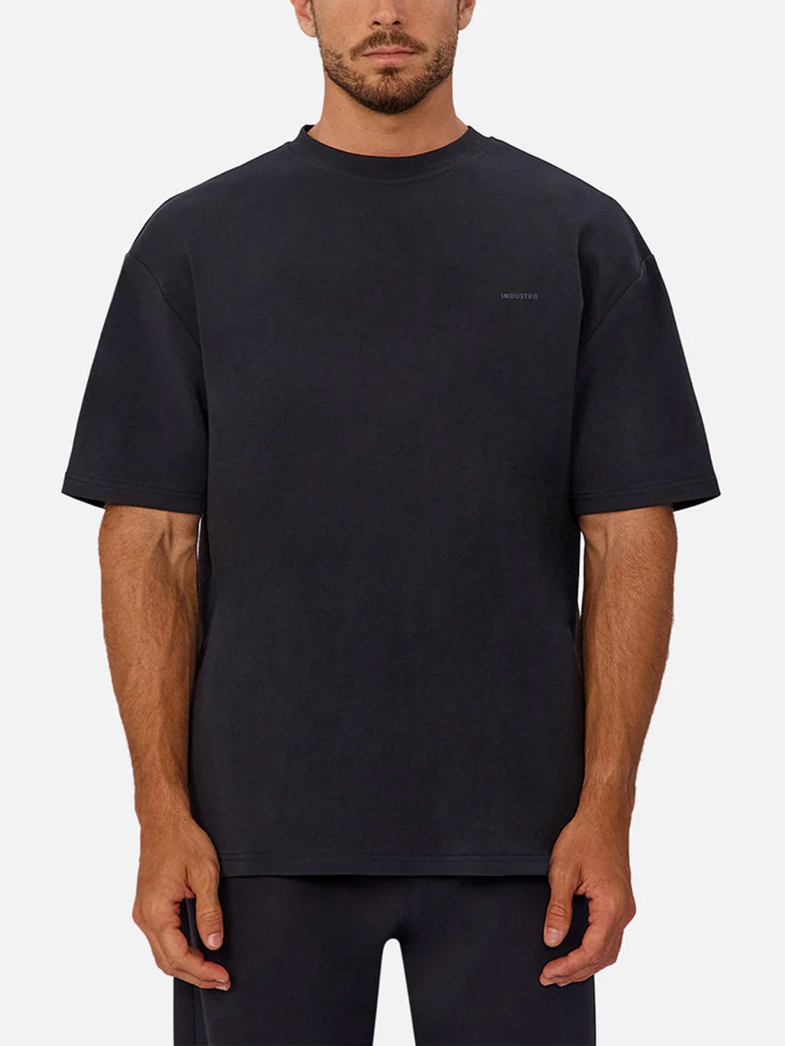 A man stands facing forward against a light gray background, wearing Industrie’s The Newport T-Shirt—a loose-fitting, heavyweight black tee with short sleeves, an oversized fit, and a small chest logo—paired with black pants.