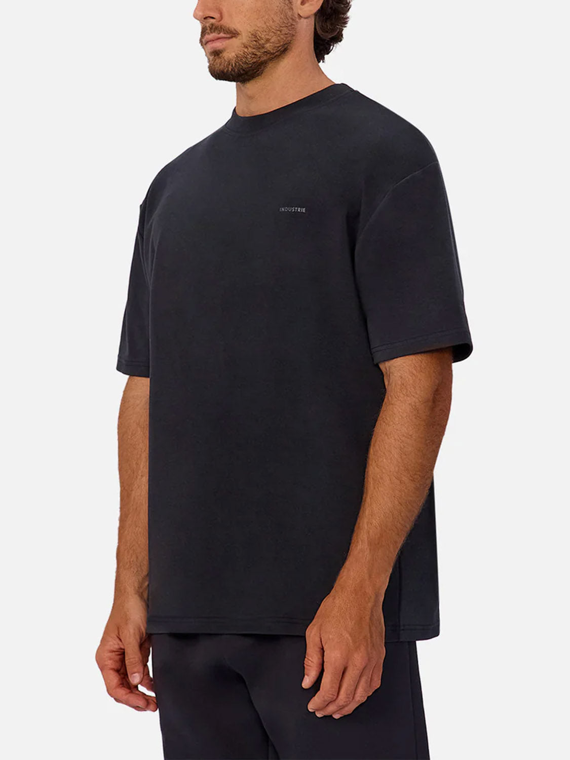 A man wears The Newport T-Shirt by Industrie, a loose-fitting, short-sleeved heavyweight tee with small white chest text, standing against a plain white background and facing slightly to the side.