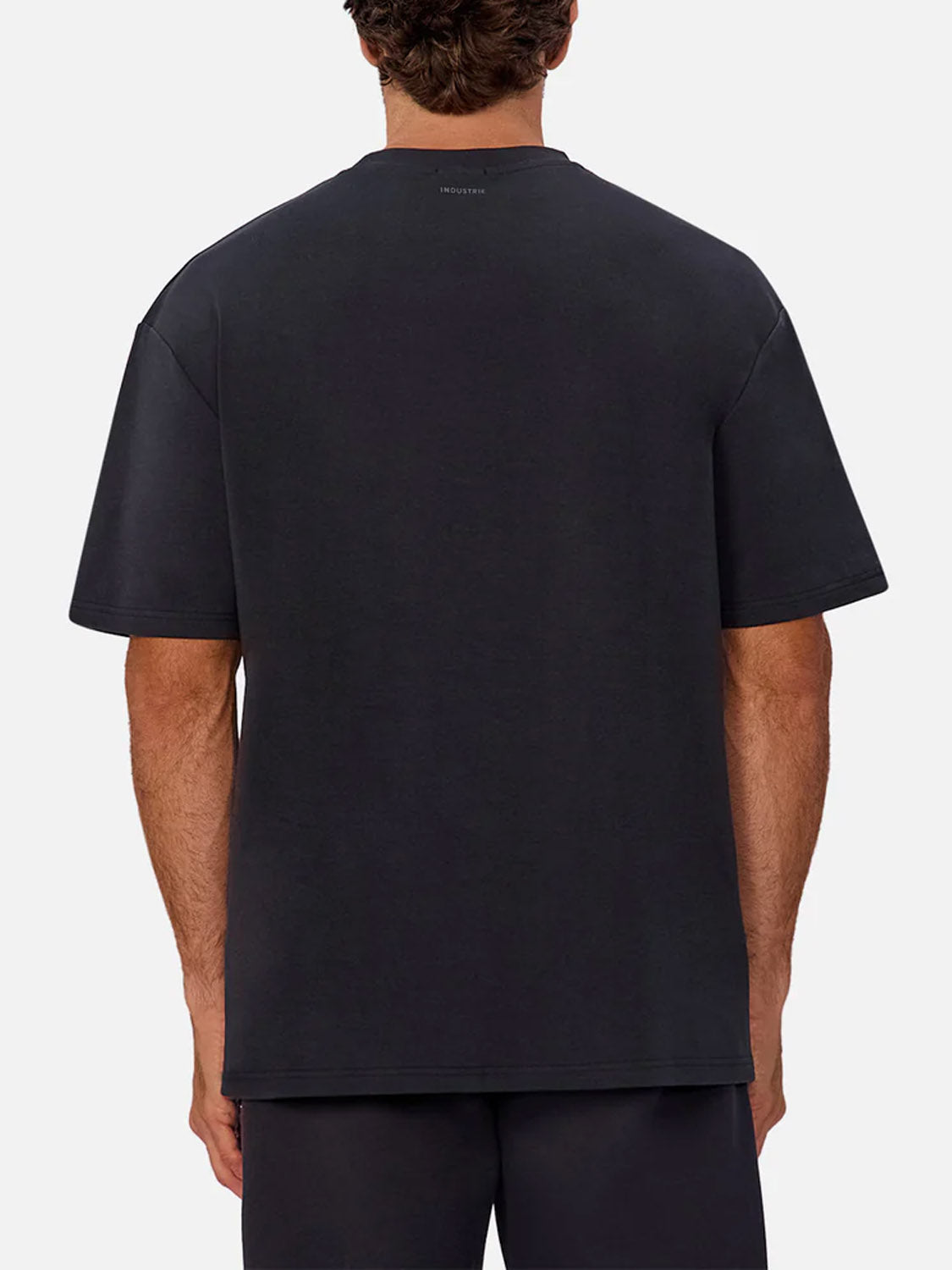 A man is seen from behind in a plain, light setting, wearing Industrie’s The Newport T-Shirt—a 90s-inspired heavyweight tee with short sleeves, an oversized relaxed fit—and dark shorts.