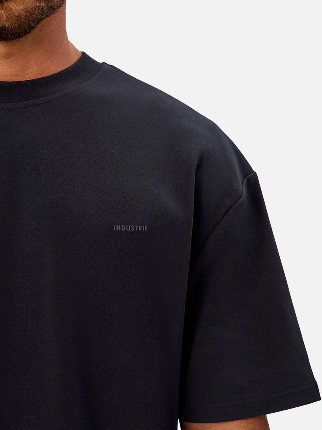 A close-up of a person wearing The Newport T-Shirt by Industrie, a plain black heavyweight tee with "Industrie" subtly printed on the left chest. The background is light gray, showing only part of the face, neck, and upper body.