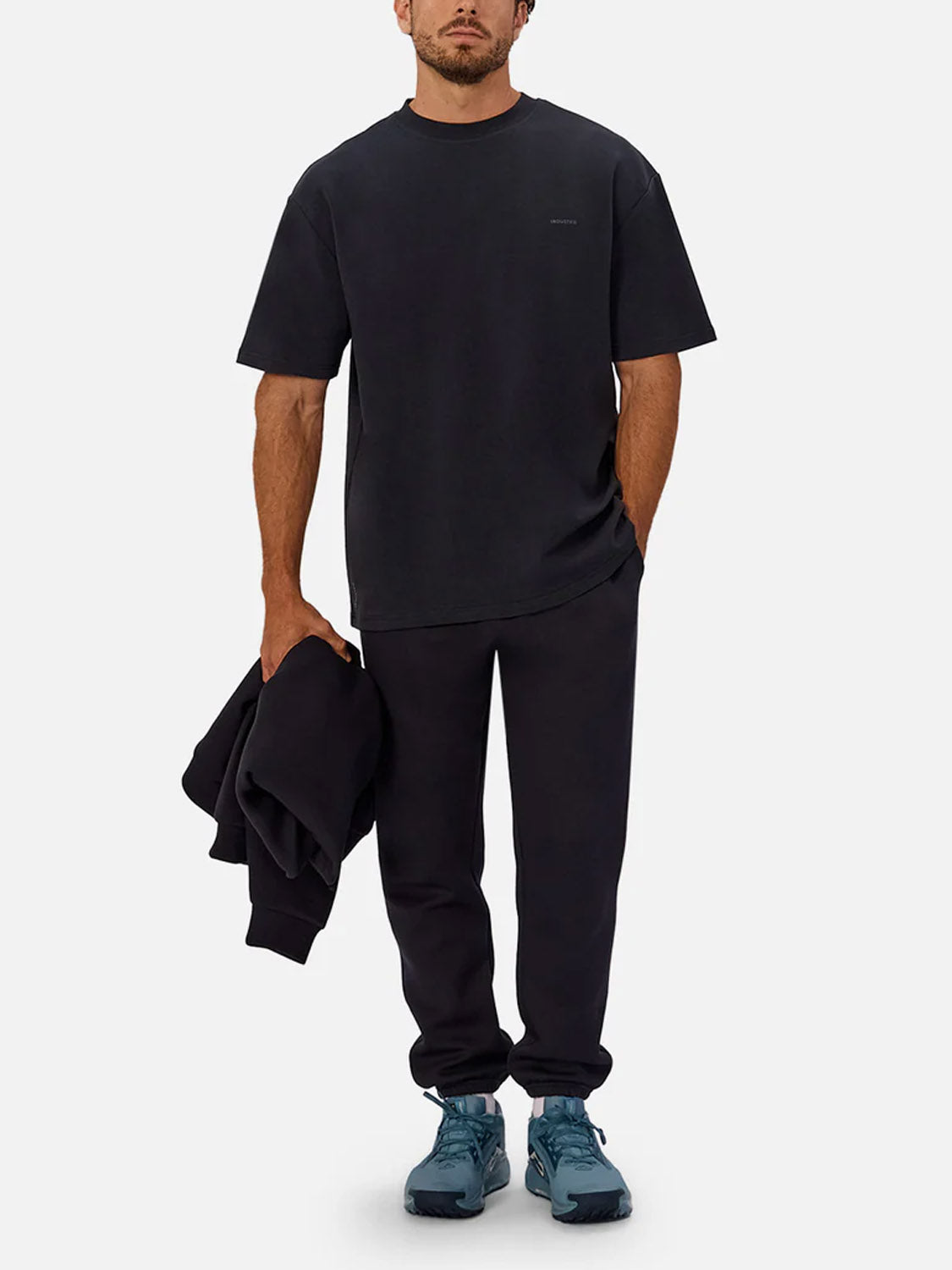 A man stands against a plain light background wearing The Newport T-Shirt by Industrie, black jogger pants, and blue sneakers, holding a black sweatshirt in one hand.