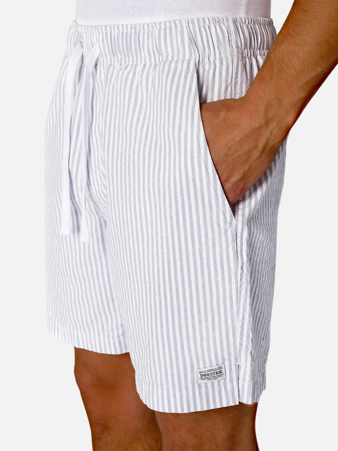 A person wearing The New Southwest Short by Industrie—men's summer shorts in light gray and white vertical stripes, with a drawstring waist, hand in pocket, and a small rectangular label near the hem. Made from 100% linen.