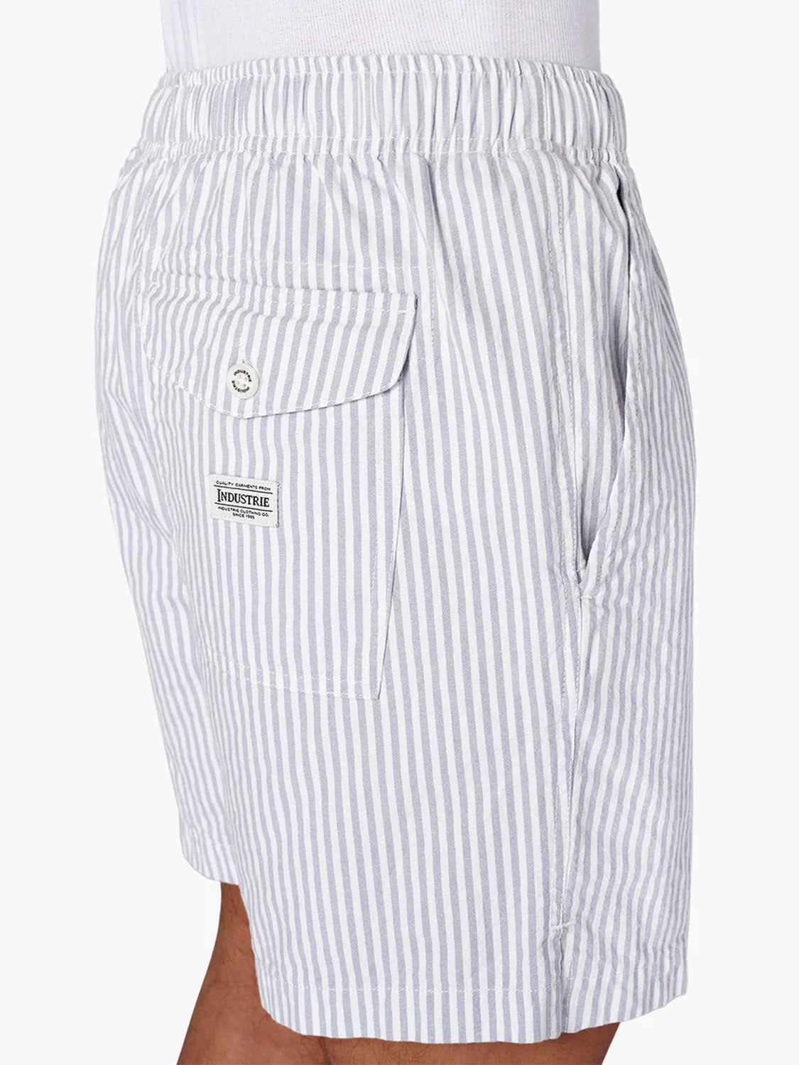 A person wearing Industrie’s The New Southwest Short—light gray and white striped 100% linen men’s summer shorts with an elastic waist, side pocket, and buttoned back pocket featuring a rectangular Huckstire Industries label.