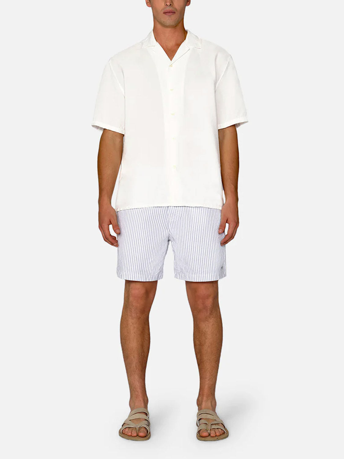 A man stands facing forward wearing Industrie’s The New Southwest Short, a light blue and white vertically striped men’s summer short, paired with a white short-sleeve button-up shirt and beige sandals against a light gray background.