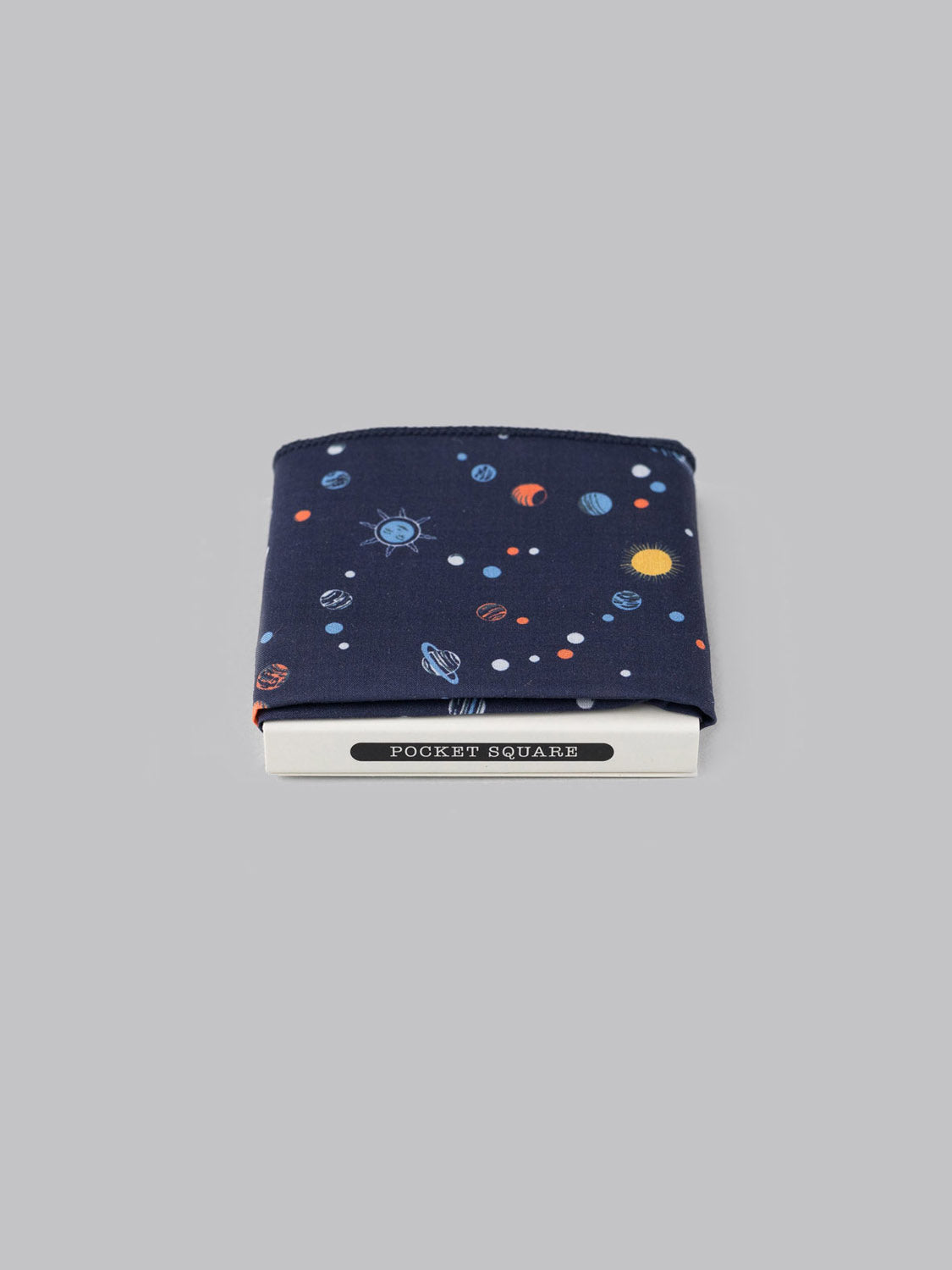 The Liberty™ Niko Pocket Square by ID Menswear features soft Tana Lawn cotton in navy blue with a vibrant space and planets design, neatly folded in white POCKET SQUARE packaging, displayed on a plain gray background.
