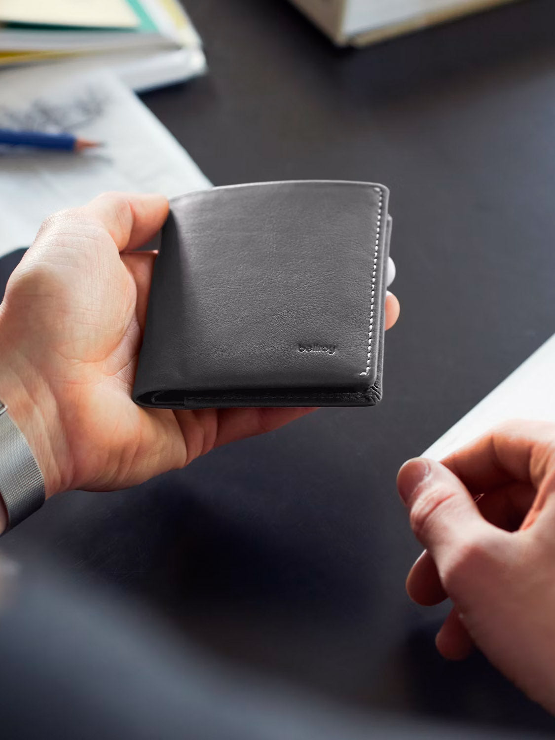 Someone holds a closed, dark gray Bellroy Note Sleeve Wallet in one hand, with the other hand nearby. Papers, a pen, and a book rest on a dark desk in the background.