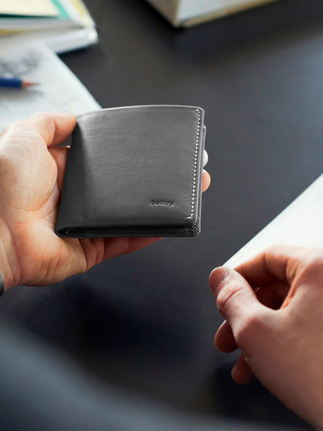 Someone holds the closed Note Sleeve Innovera™ Edition wallet by Bellroy, in black leather alternative with the brand name embossed on front, while seated at a desk with papers and a pen in the background.