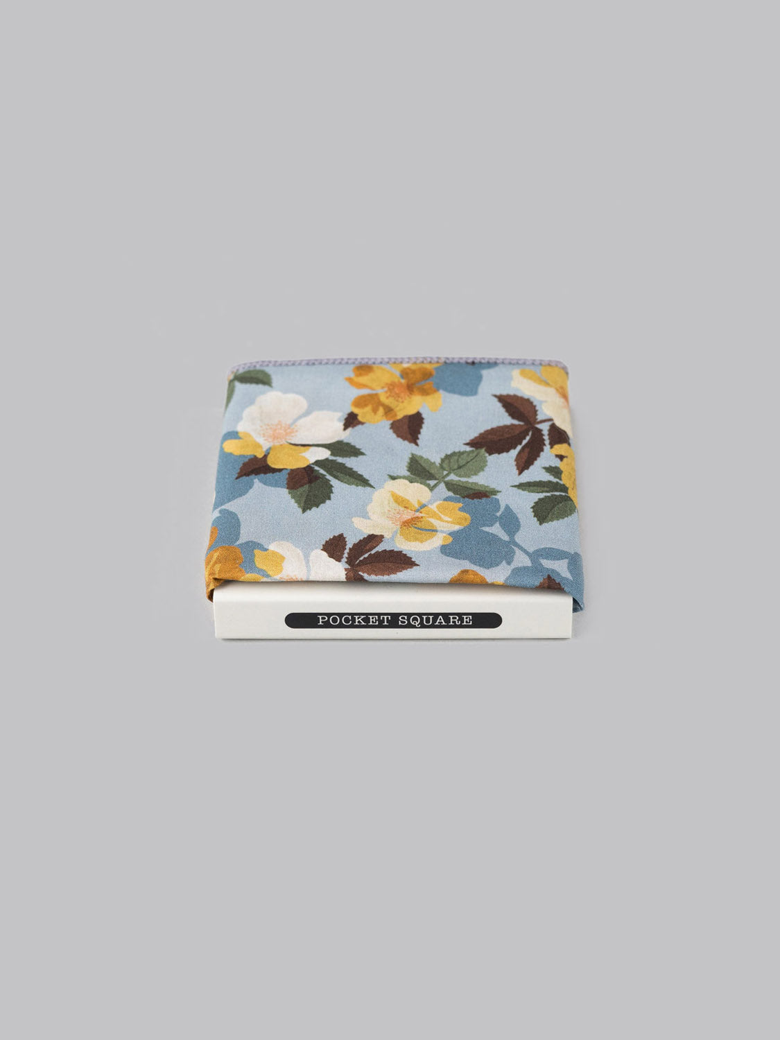The Liberty™ Nysa Pocket Square by ID Menswear, handmade in Brooklyn from floral light blue Tana Lawn cotton with yellow, white, and green accents, is displayed folded atop a POCKET SQUARE box on a plain gray background.