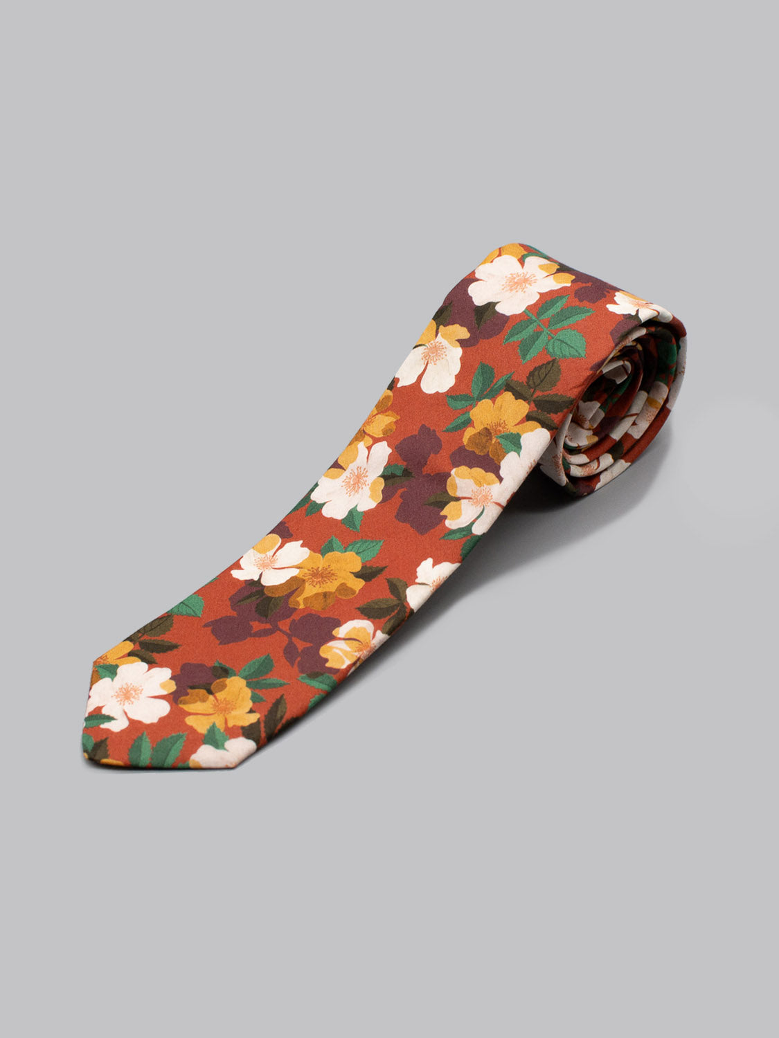 The Liberty of London Tana Lawn™ Nysa Tie by ID Menswear features a handmade floral pattern in white, yellow, and green on a reddish-brown background, crafted from premium Tana Lawn™ fabric and displayed on a plain gray surface.