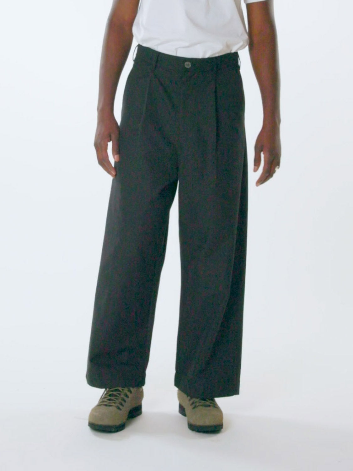 A person stands against a plain background wearing Obey Oliver Pleated Pants, white t-shirt, and beige lace-up shoes. The focus is on the lower half of the body, showcasing the relaxed fit and style of both the pants and footwear.