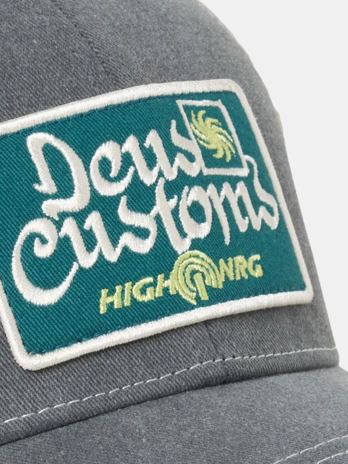 Close-up of the Deus Open Head Trucker, a gray cotton twill cap in a classic fit, featuring a green and white embroidered patch with "Deus Customs" and "HIGH NRG" plus stylized yellow designs.