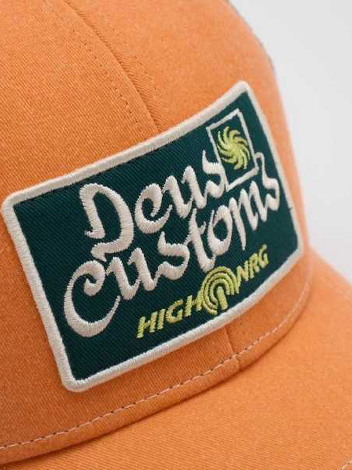 A close-up of the Deus Open Head Trucker, an orange cotton twill cap with a green and white embroidered patch reading "Deus Customs HIGH NRG" and a yellow spiral graphic.