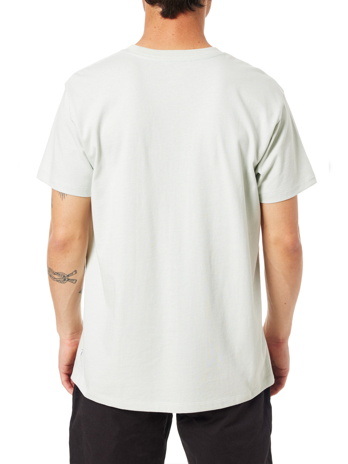 A person with short hair wears the Katin Orb Embroidered Tee by Katin and black pants, shown from behind on a white background. Their visible arms include a tattoo on the left forearm.