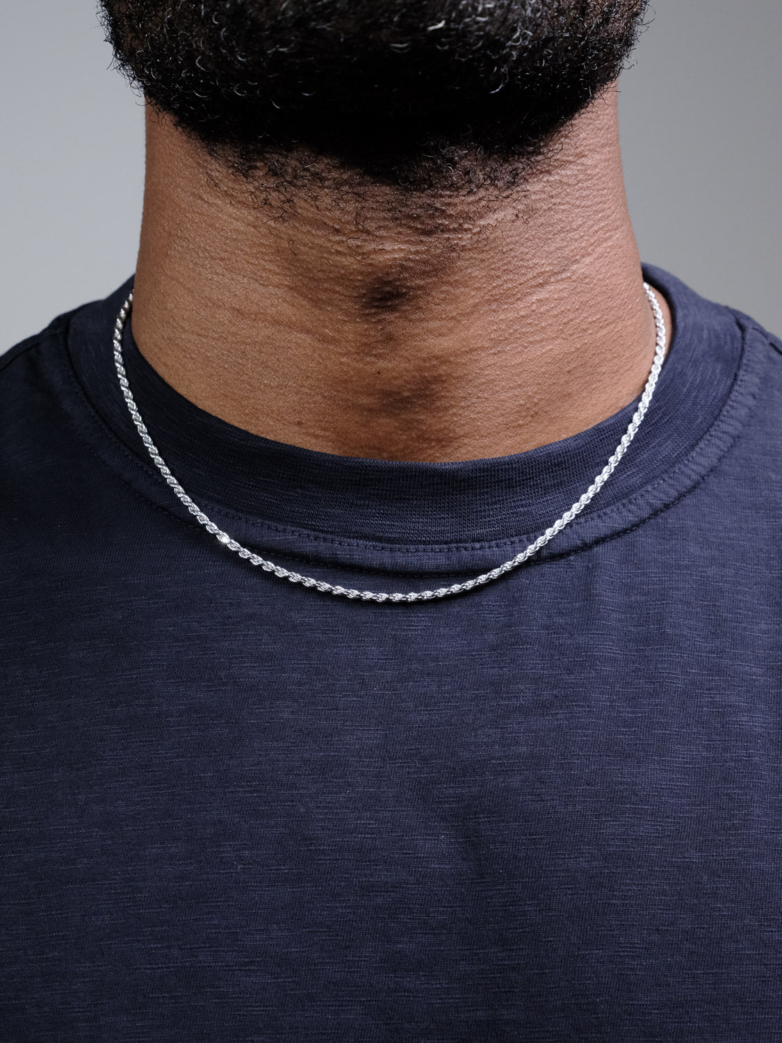 A close-up of a neck and upper chest showcases the Orion 50 Rope Chain Necklace by ID Menswear, a 4mm sterling silver piece made in Italy. Worn with a dark blue crew-neck shirt; face not shown against a plain light gray background.