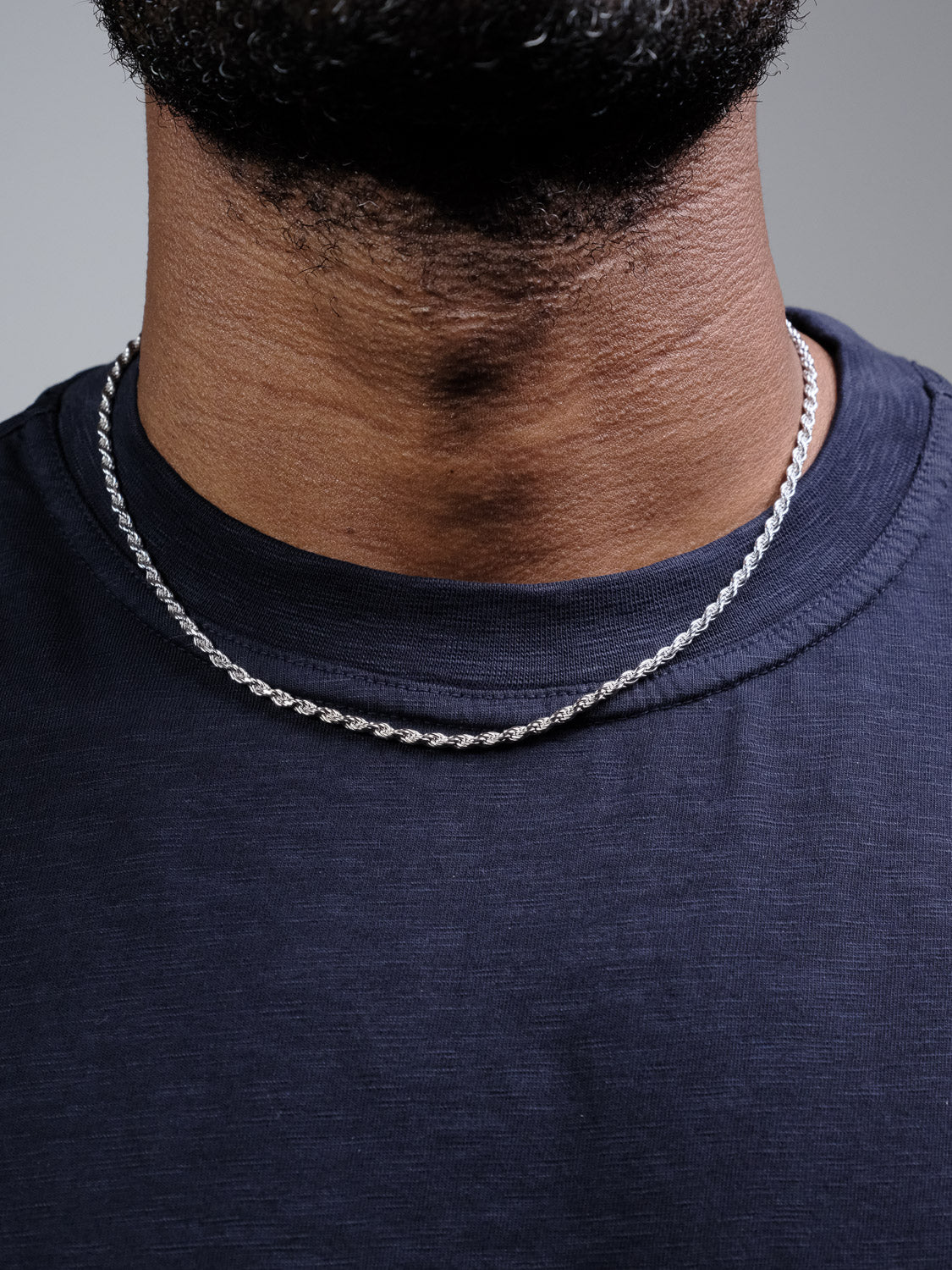 A close-up of a person's neck wearing the Orion 60 Rope Chain Necklace by ID Menswear, crafted from 925 Sterling Silver in Italy. The necklace stands out against a plain background and pairs with a dark navy blue crew-neck shirt.