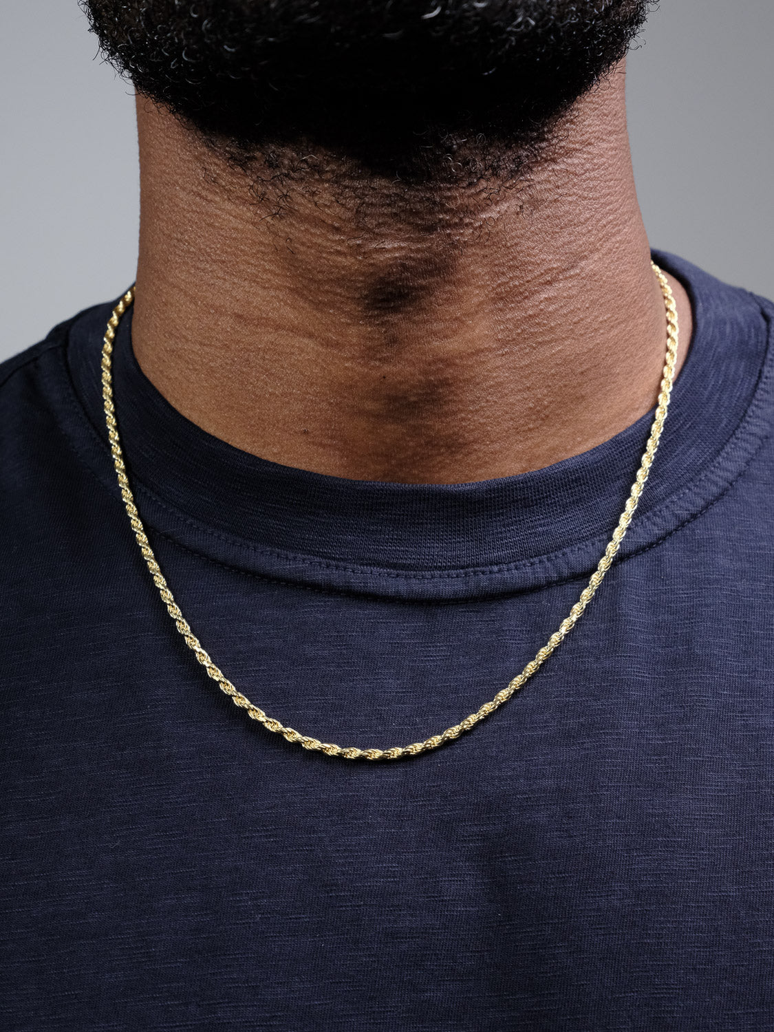 A close-up shot shows a person wearing an ID Menswear Orion 60 Rope Chain Necklace, gold plated over 925 Sterling Silver, along with a navy blue shirt. Only their lower face and neck are visible.