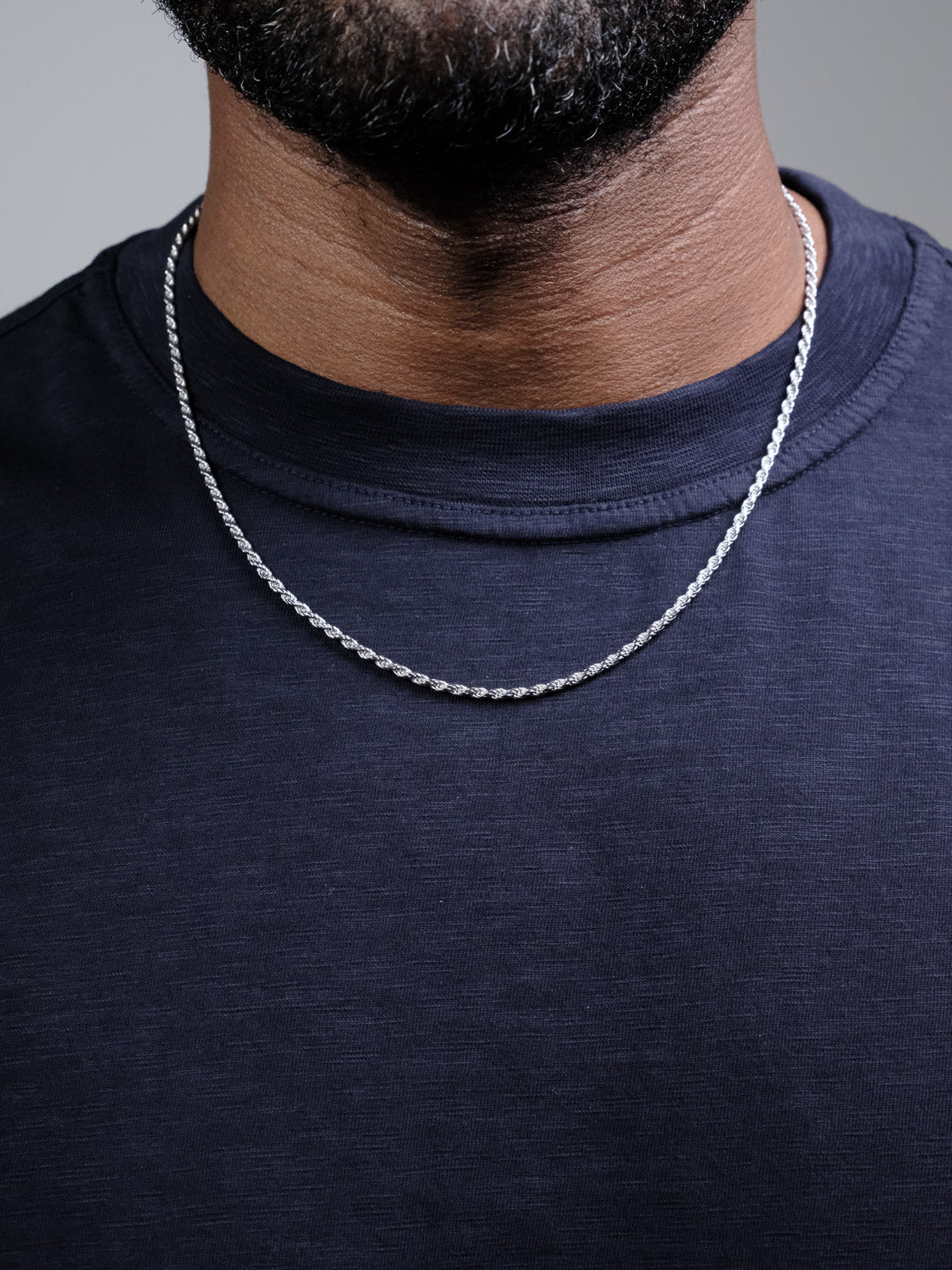 A close-up shows a person wearing a dark navy shirt and the Orion 50 Rope Chain Necklace by ID Menswear, a sleek 4mm sterling silver piece visible on their neck and upper chest. Only the lower face is shown.
