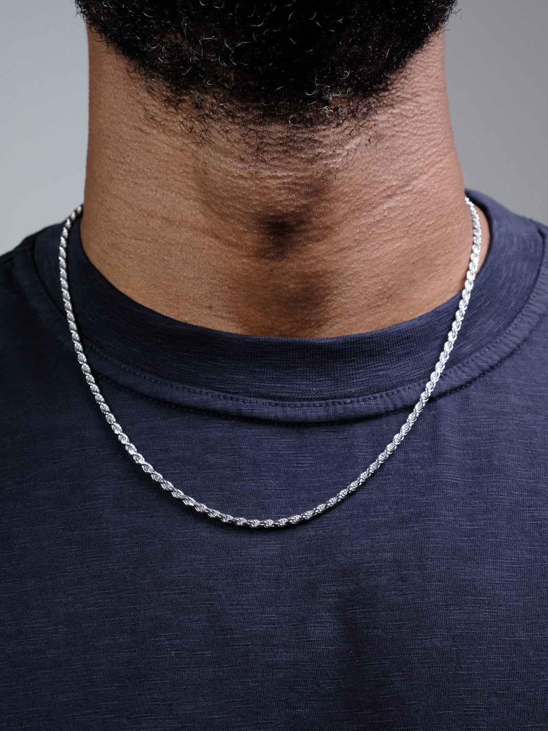 A close-up of a person wearing the Orion 60 Rope Chain Necklace by ID Menswear, paired with a dark blue crew-neck shirt, showing only the lower face and neck.