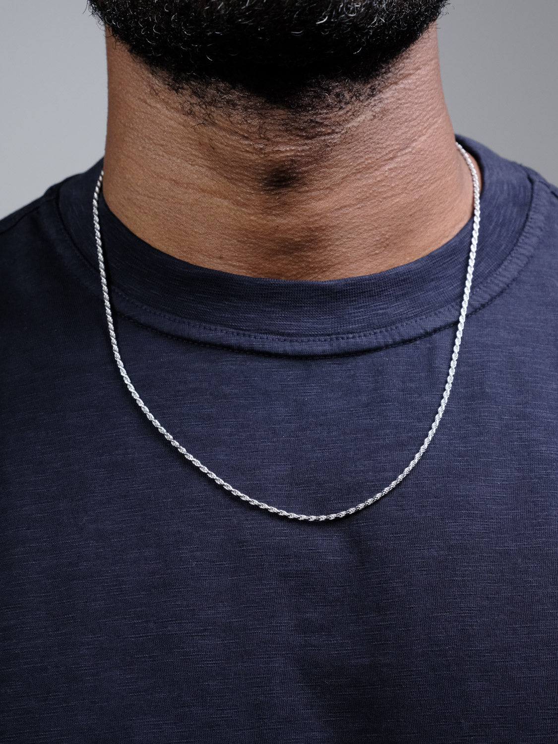 A close-up shows a person wearing the Orion 40 Rope Chain Necklace by ID Menswear over a dark navy t-shirt. The lower face and neck are visible against a neutral background.